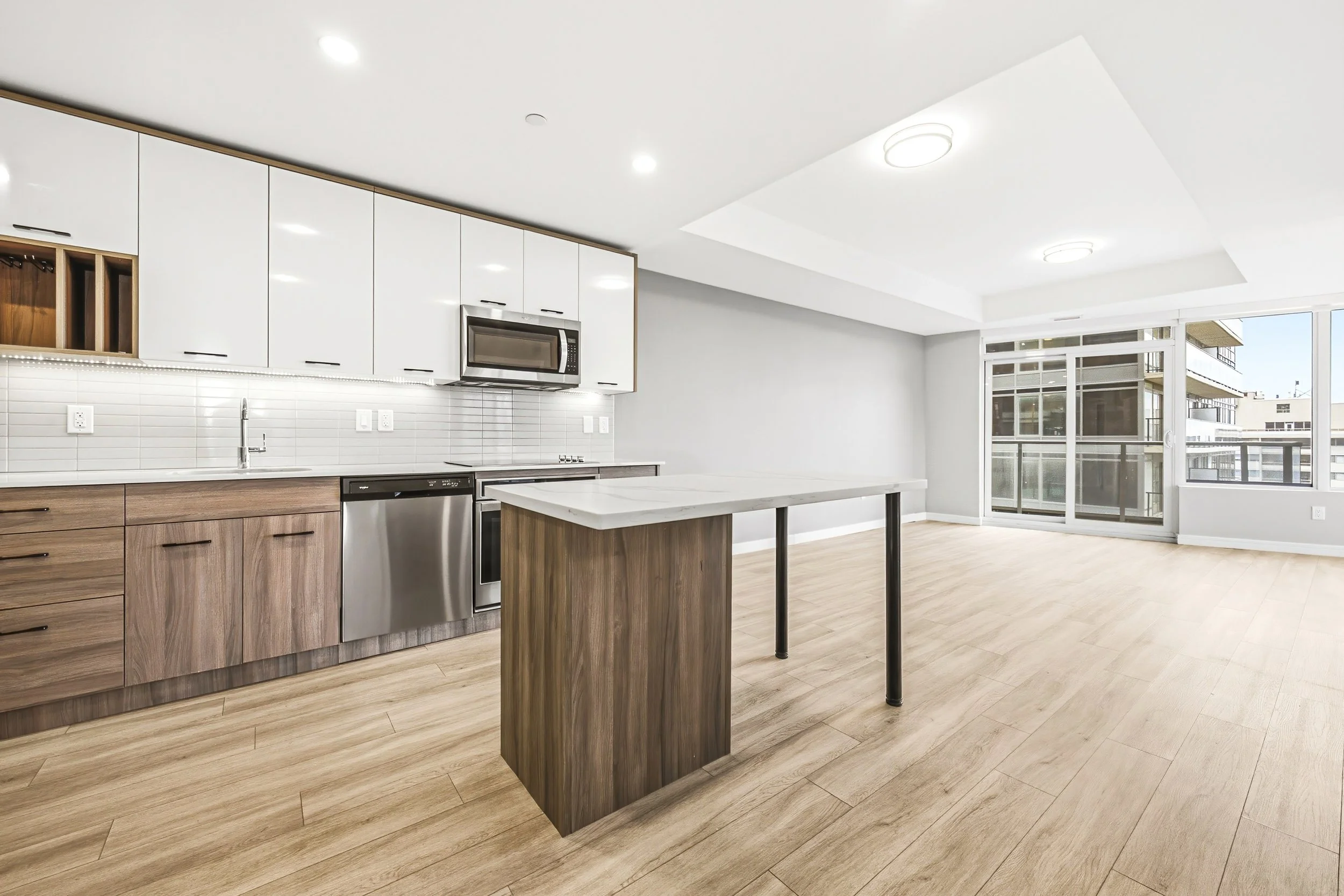 Modern kitchen with wooden cabinets, white upper cabinets, stainless steel appliances, a kitchen island, wood flooring, large windows, and balcony doors.