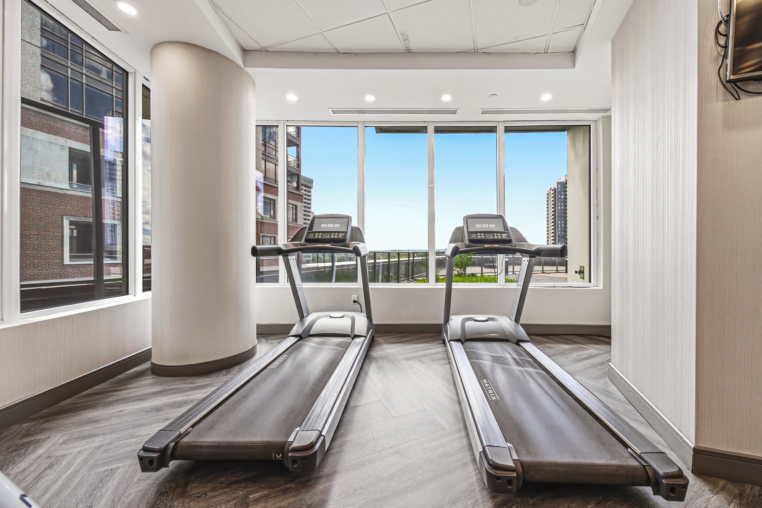 Two treadmill desks facing large windows in a modern apartment or gym, with buildings and blue sky outside.