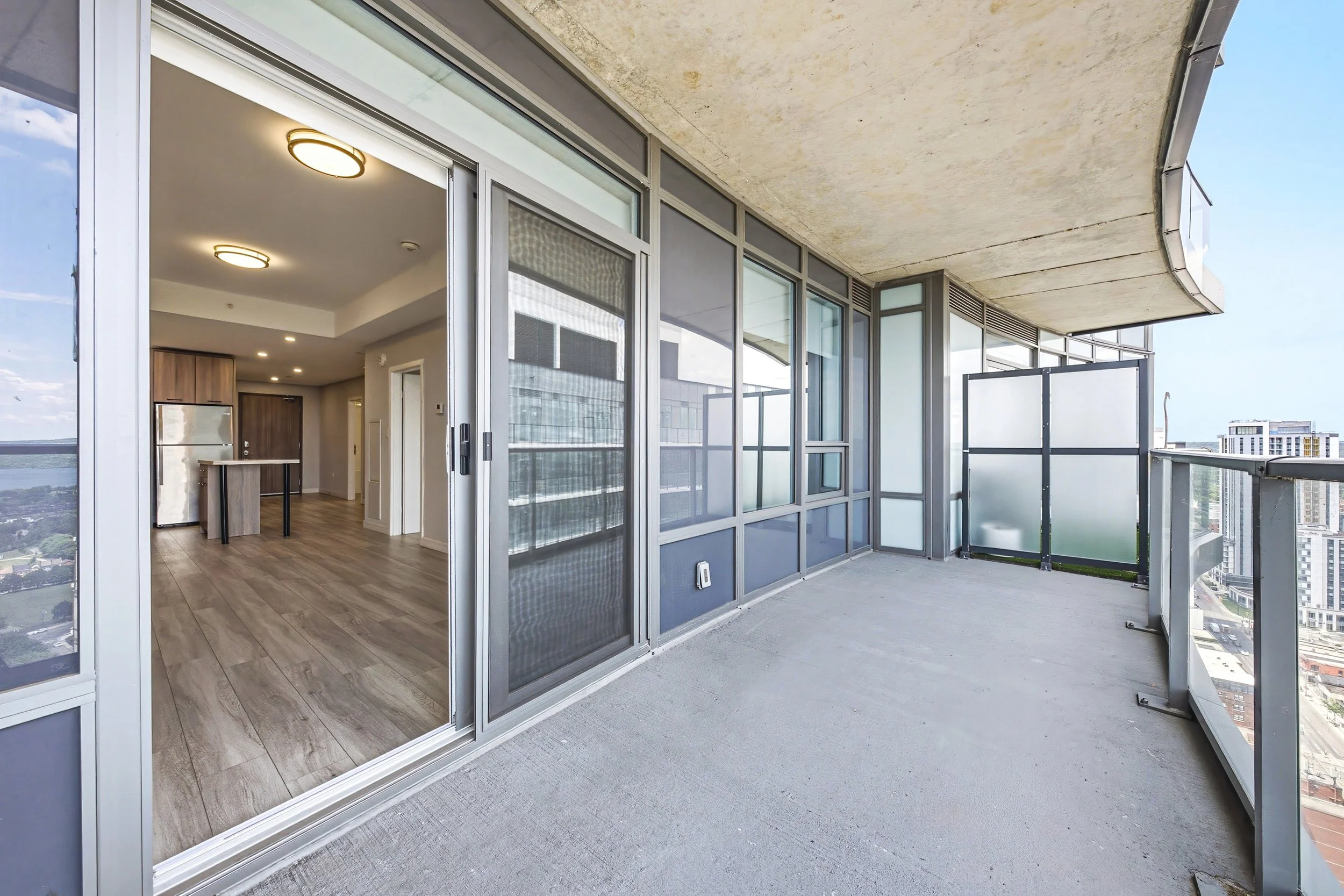 View of a modern apartment balcony with frosted glass safety railing and exterior concrete ceiling, overlooking a city with high-rise buildings.
