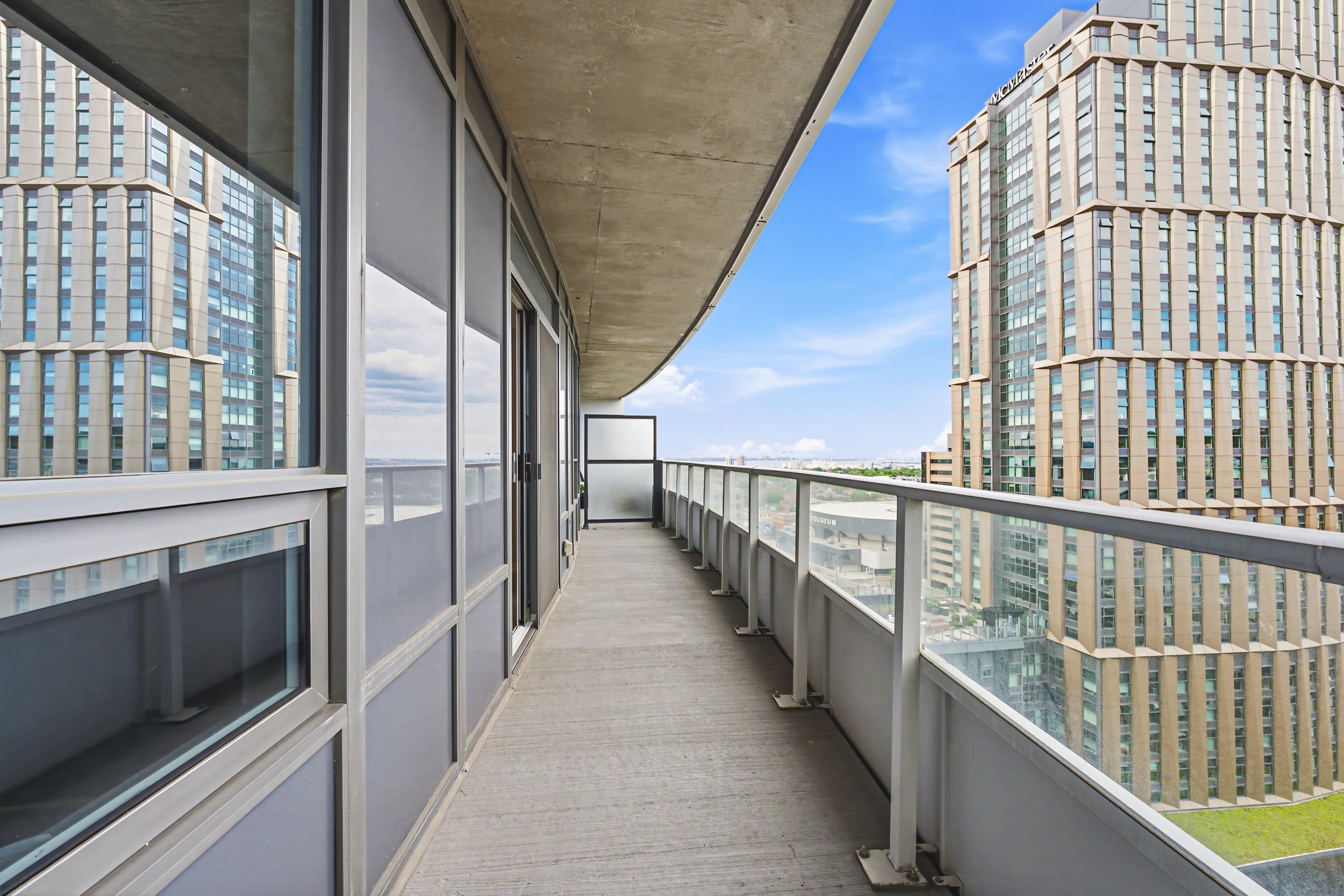 An outdoor balcony with a concrete ceiling and metal railing, overlooking tall modern skyscrapers under a partly cloudy sky.