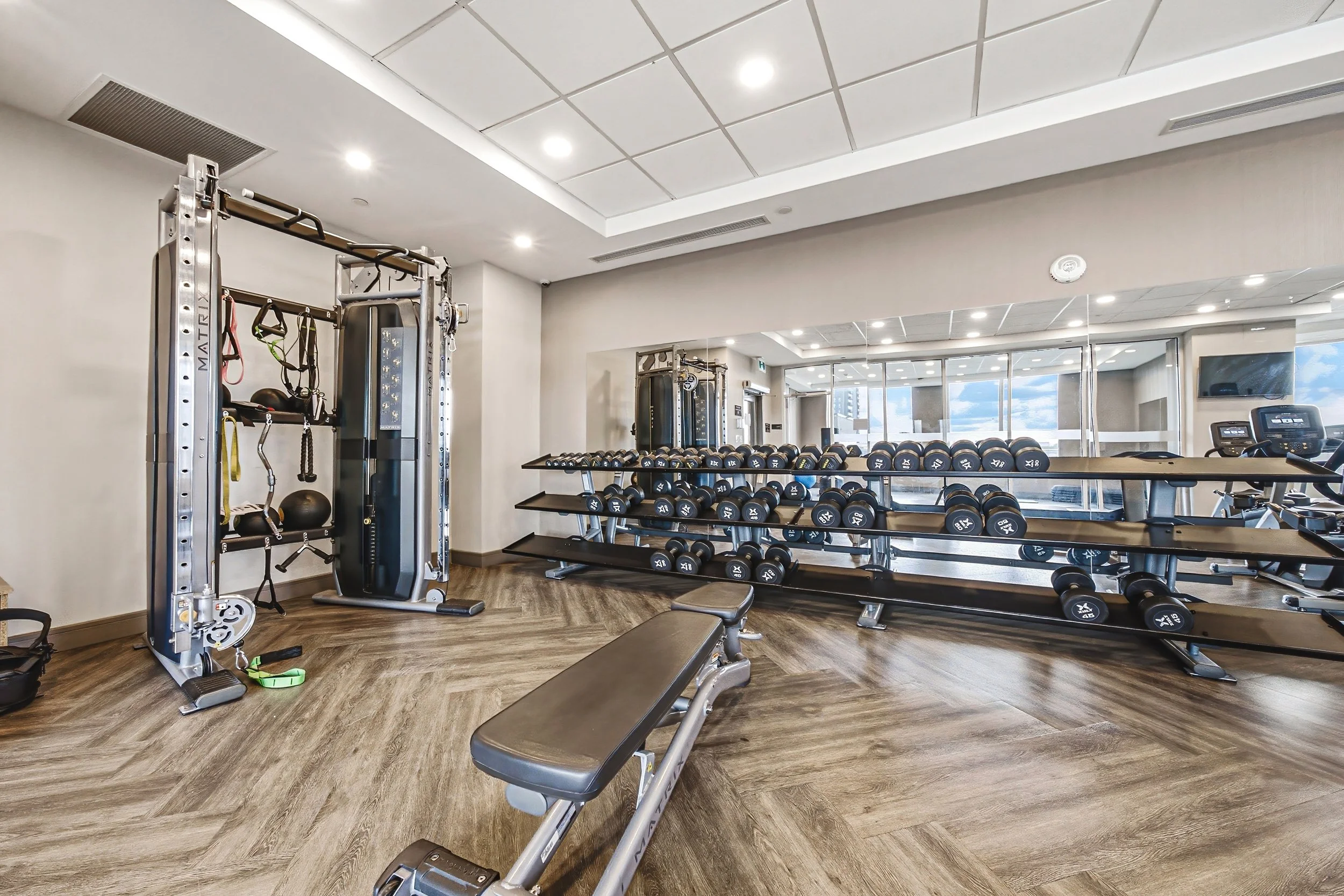 Fitness room with dumbbell rack, weight machines, benches, and large windows showing a cityscape.
