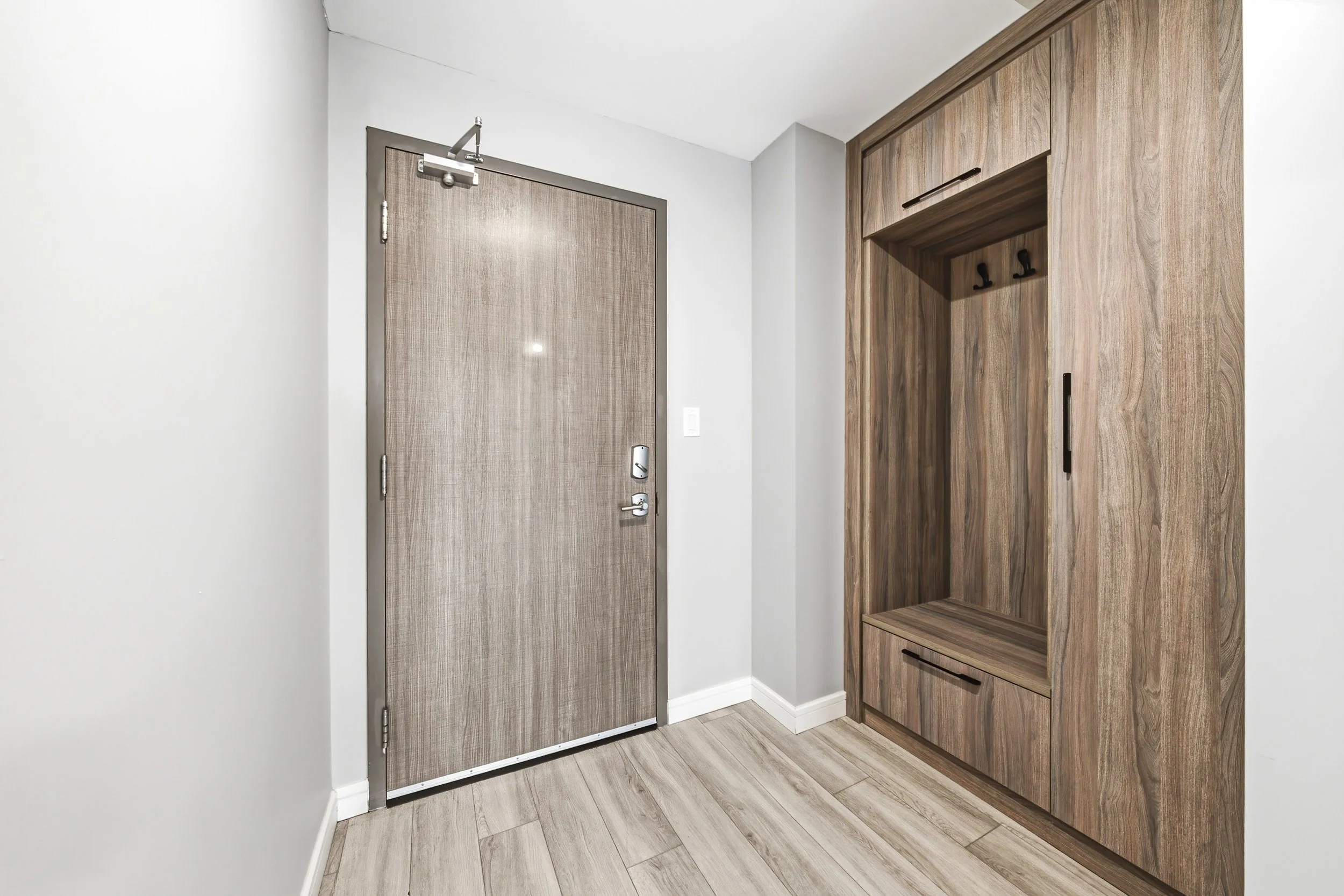 Interior view of a room with a closed wooden door on the left and a wooden built-in cabinet on the right, with coat hooks and drawers, and light-colored flooring.