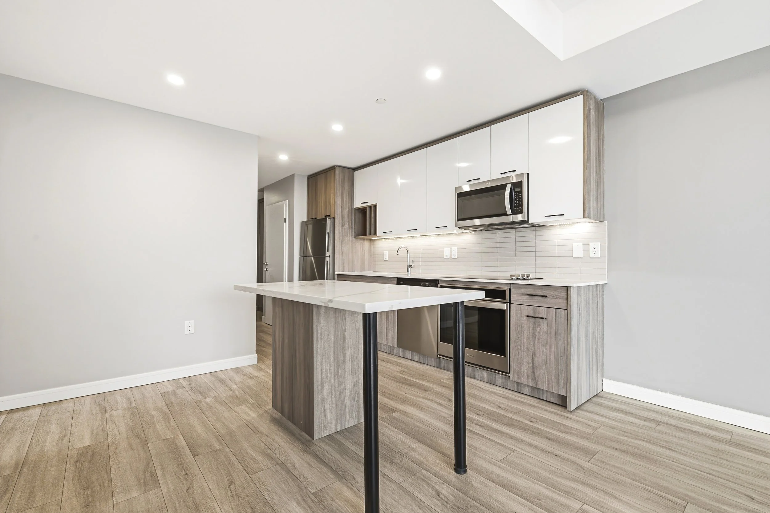 Modern kitchen with white upper cabinets, wooden lower cabinets, stainless steel appliances including a microwave, American refrigerator, and oven, with a white countertop and light wood flooring.