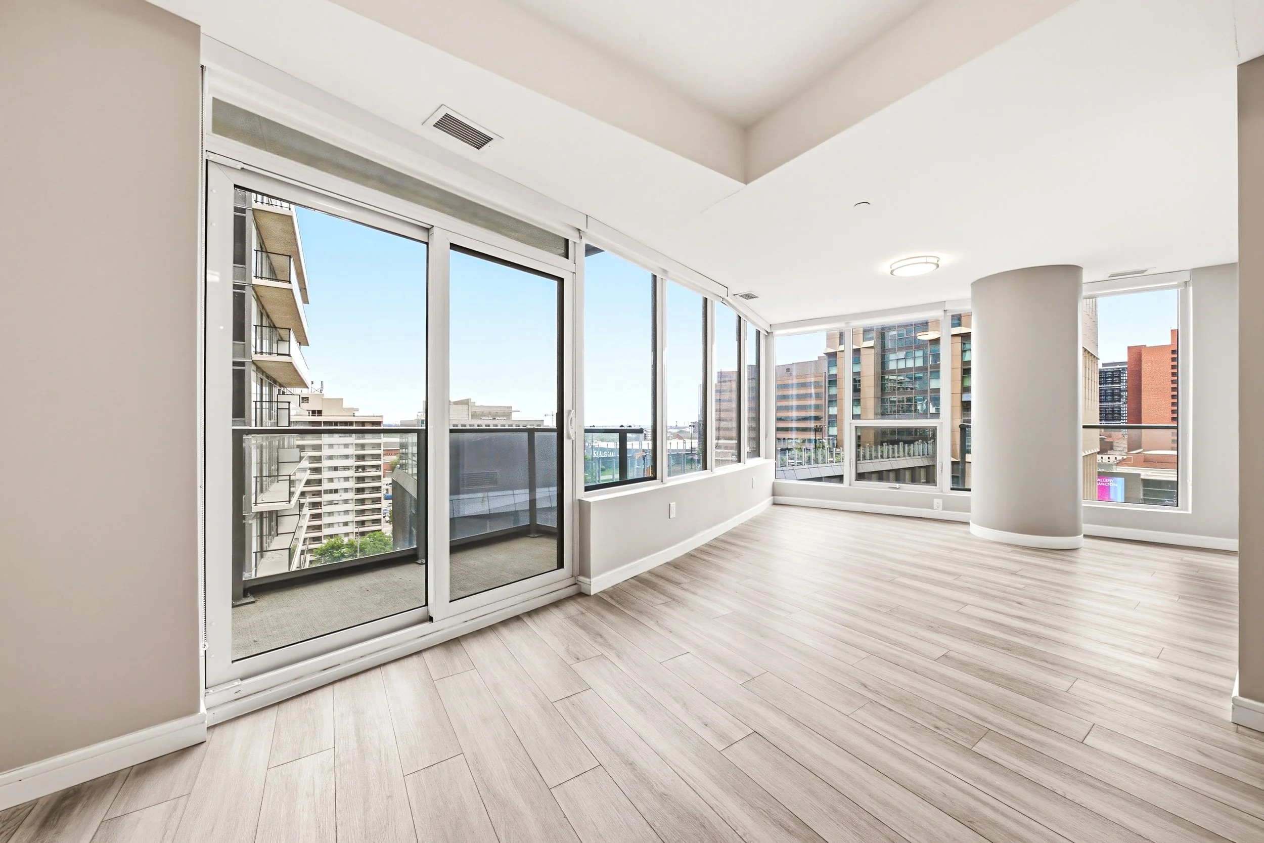 Empty high-rise apartment room with large floor-to-ceiling windows showing city view, light wood flooring, white ceiling and walls, and a white column.