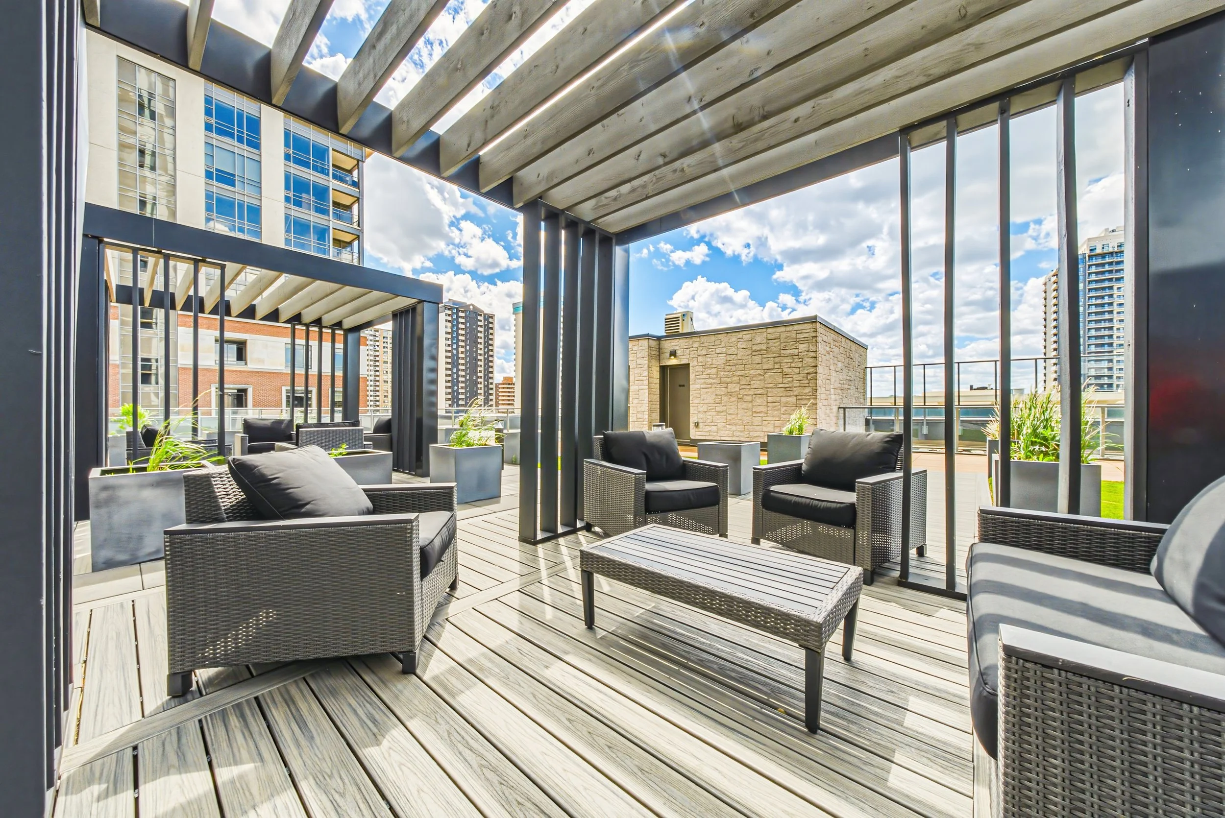 Outdoor rooftop patio with black wicker chairs and coffee tables, surrounded by modern buildings against a bright, partly cloudy sky.