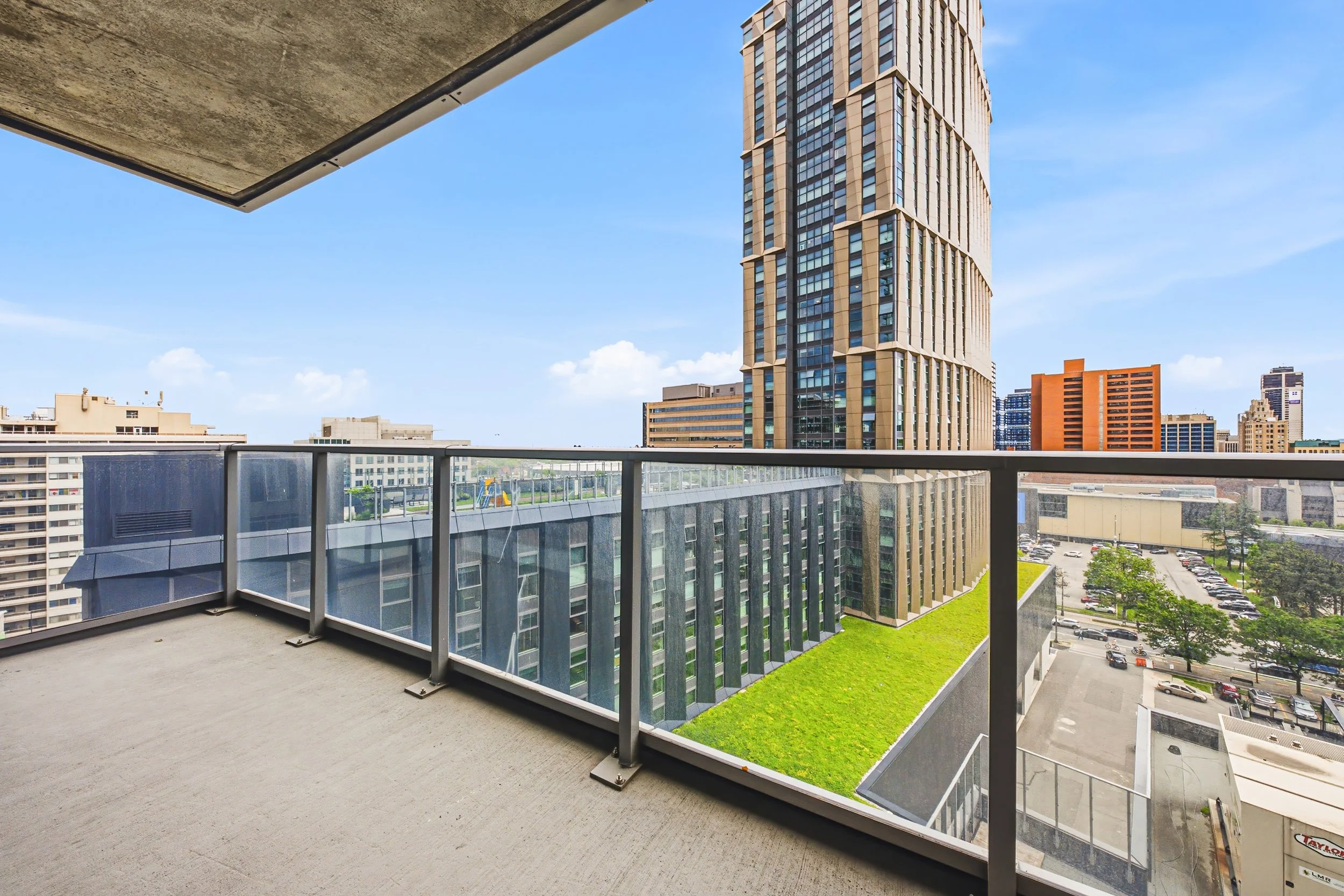 View from a balcony of a modern urban high-rise building, showing the rooftop of a lower building with a glass railing, and a cityscape of tall buildings against a blue sky.