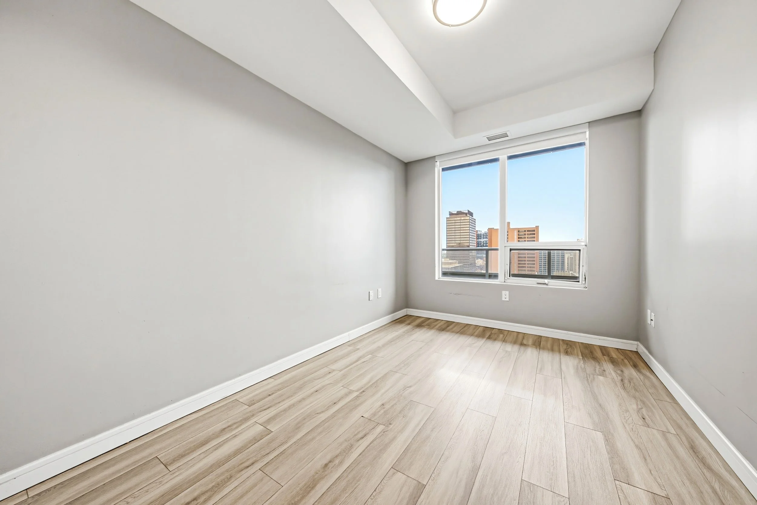 Empty room with light wood flooring, white walls, large window with city view, and upper ceiling light fixture.