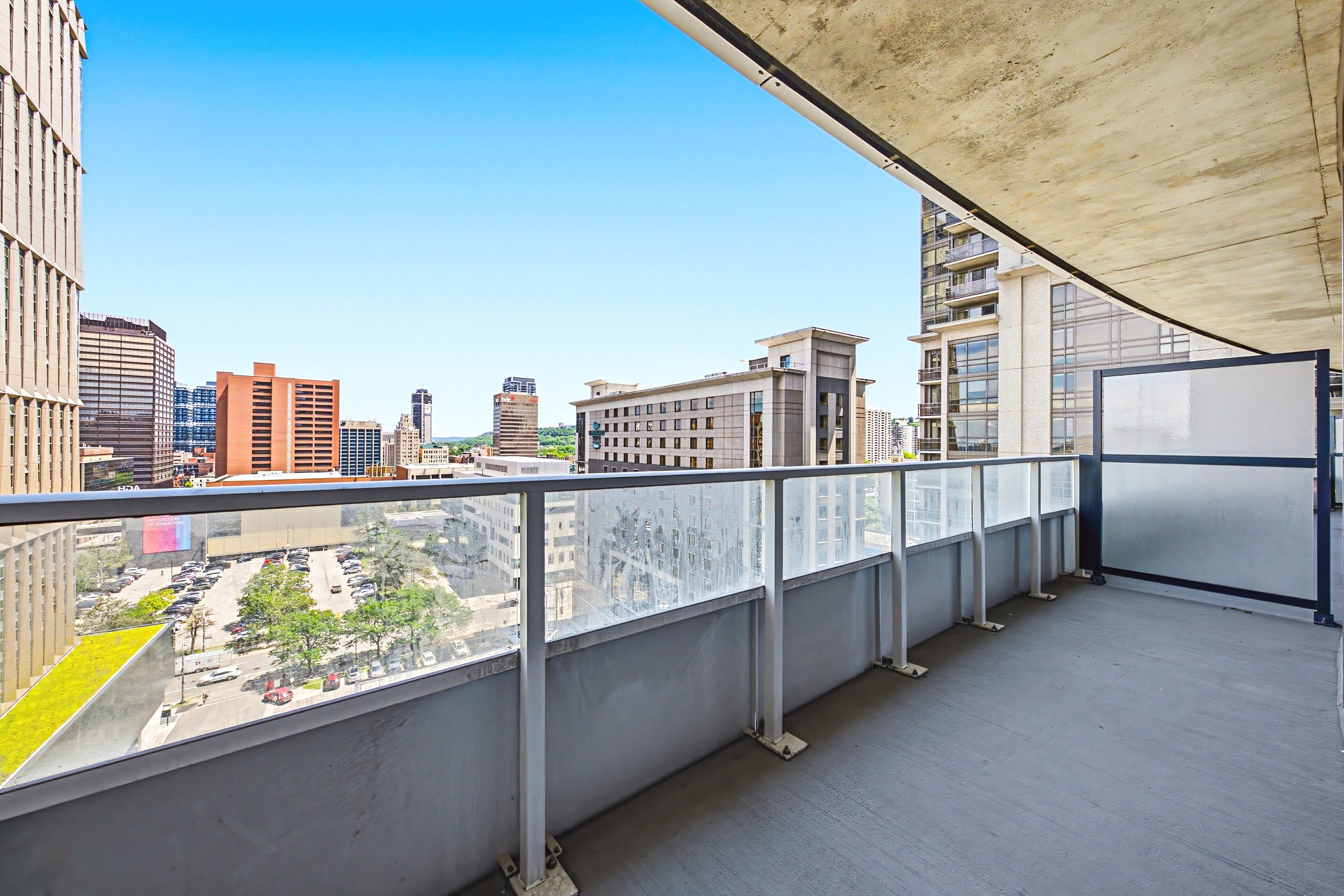 View from a balcony overlooking a cityscape with tall buildings, parking lot, and trees under a clear blue sky.