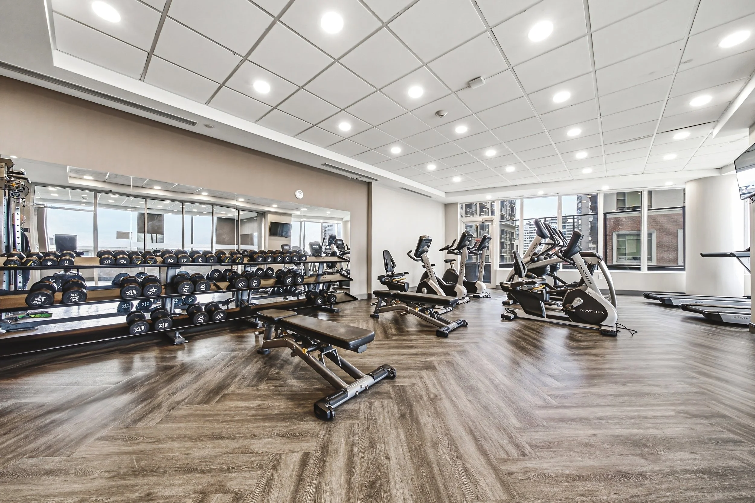 A spacious gym room with treadmills, elliptical machines, and a rack of dumbbells, featuring large windows, wood flooring, and a ceiling with recessed lighting.