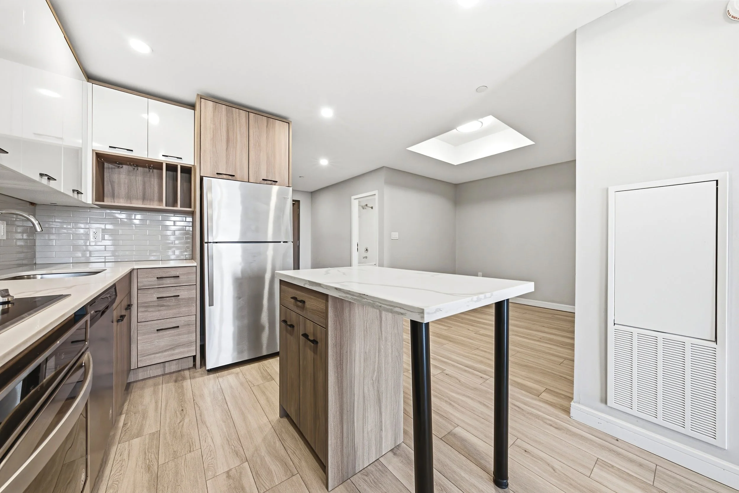 Modern kitchen with light wood cabinets, stainless steel refrigerator, white marble island, and light wood flooring, illuminated by ceiling recessed lights and a skylight.