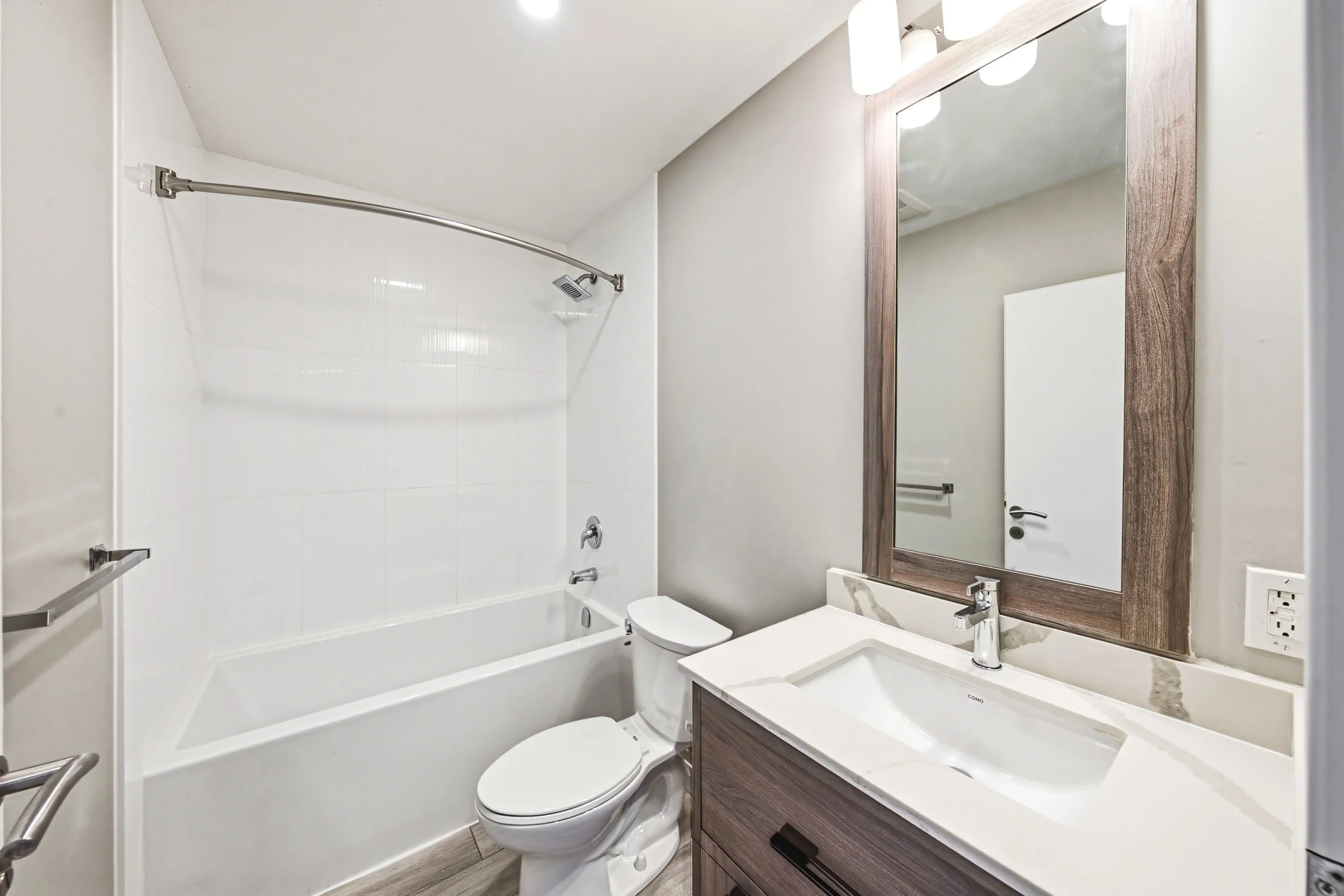 A modern bathroom with a bathtub and shower on the left, a toilet in the center, and a vanity with a sink and large mirror on the right. The walls are painted light gray, and there is a wooden frame around the mirror.