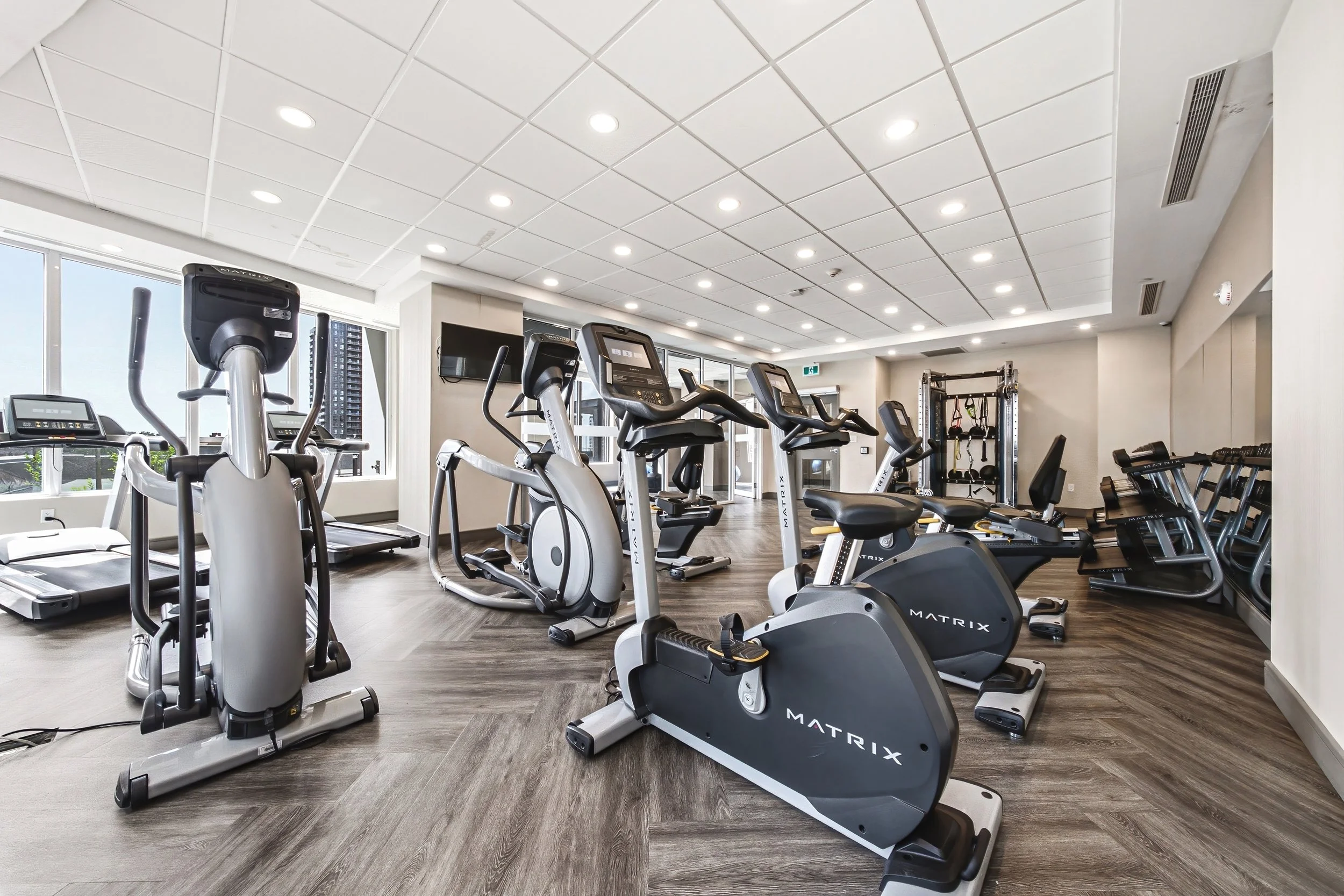 Empty gym with various cardio equipment including treadmills, stationary bikes, and elliptical machines, in a well-lit space with wood flooring and large windows.