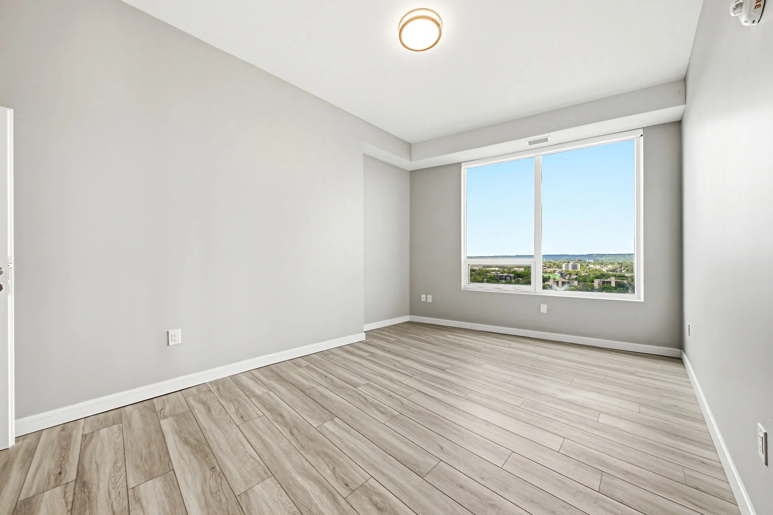 Empty modern room with large window showing a city view, white walls, wood flooring, ceiling light, and electrical outlets.