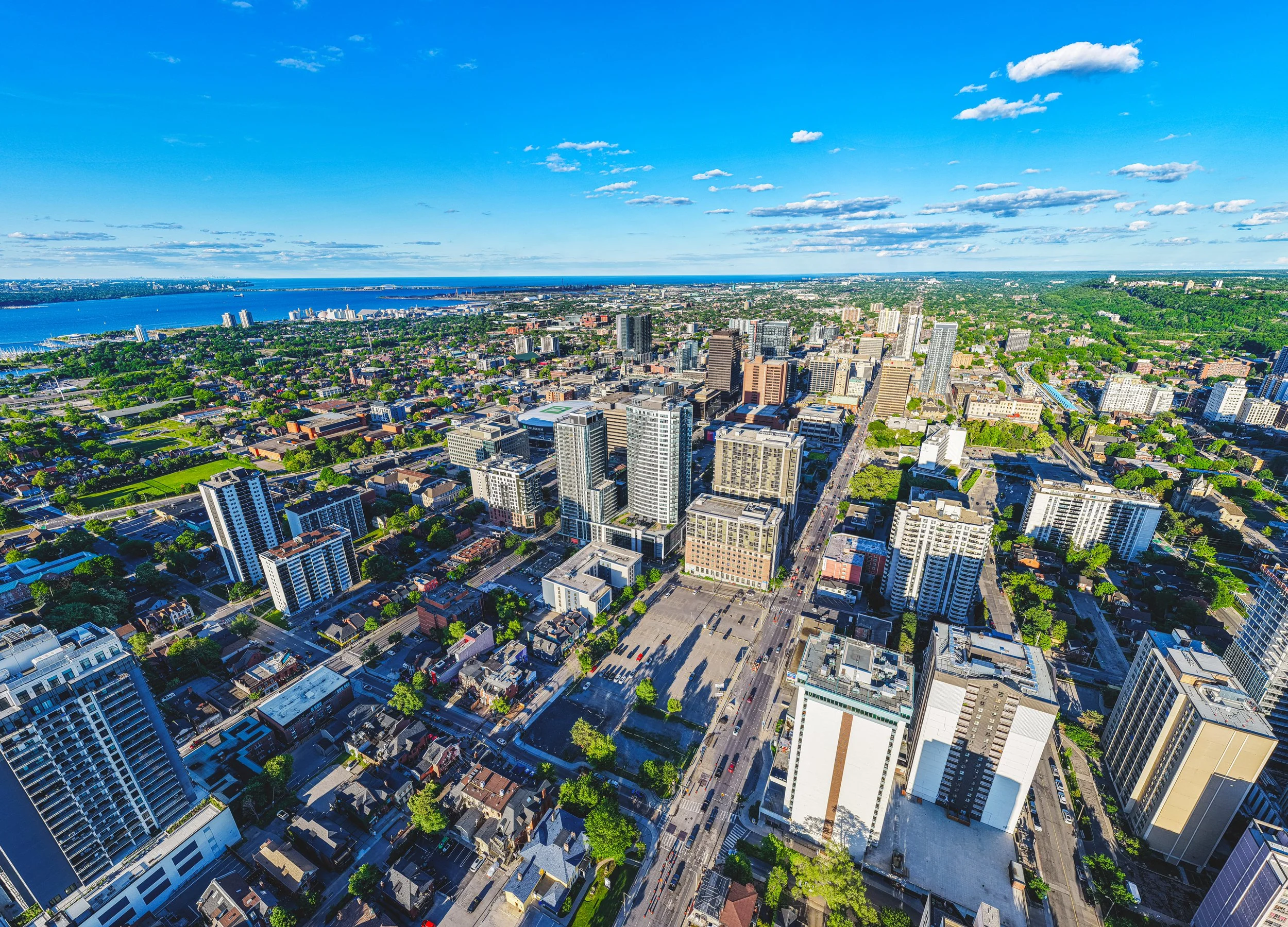 An aerial view of a cityscape during daytime, featuring numerous tall buildings, a river, and green parks under a blue sky with scattered clouds.