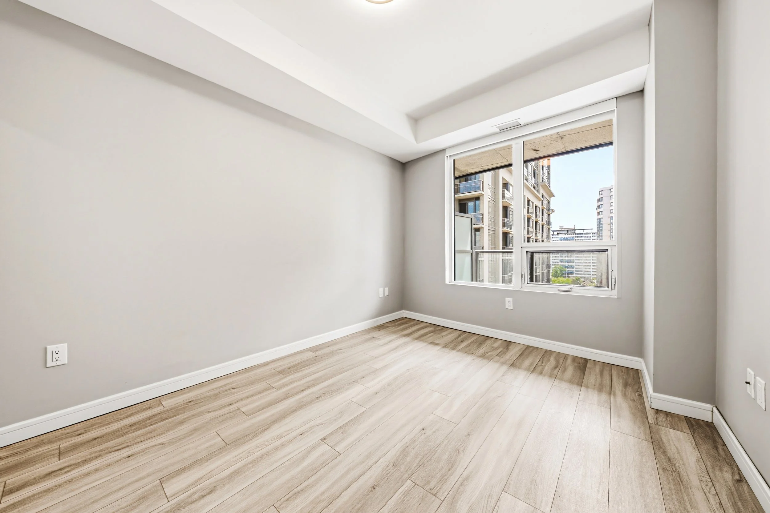 Empty room with light wood flooring, white walls, a large window showing a cityscape of apartment buildings, and several electrical outlets.