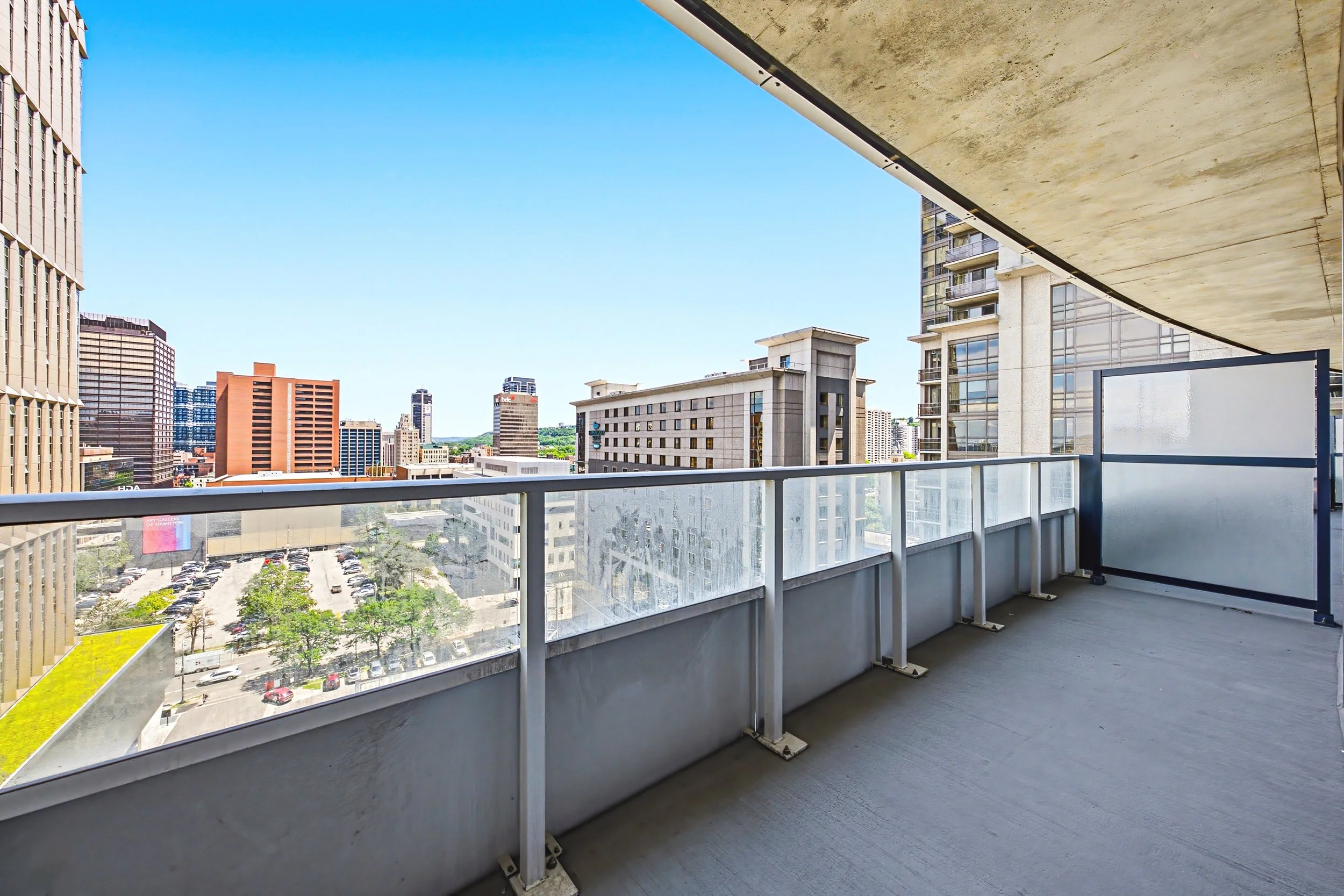 View from a balcony overlooking a city skyline with tall buildings, parking lot, and green trees under a clear blue sky.
