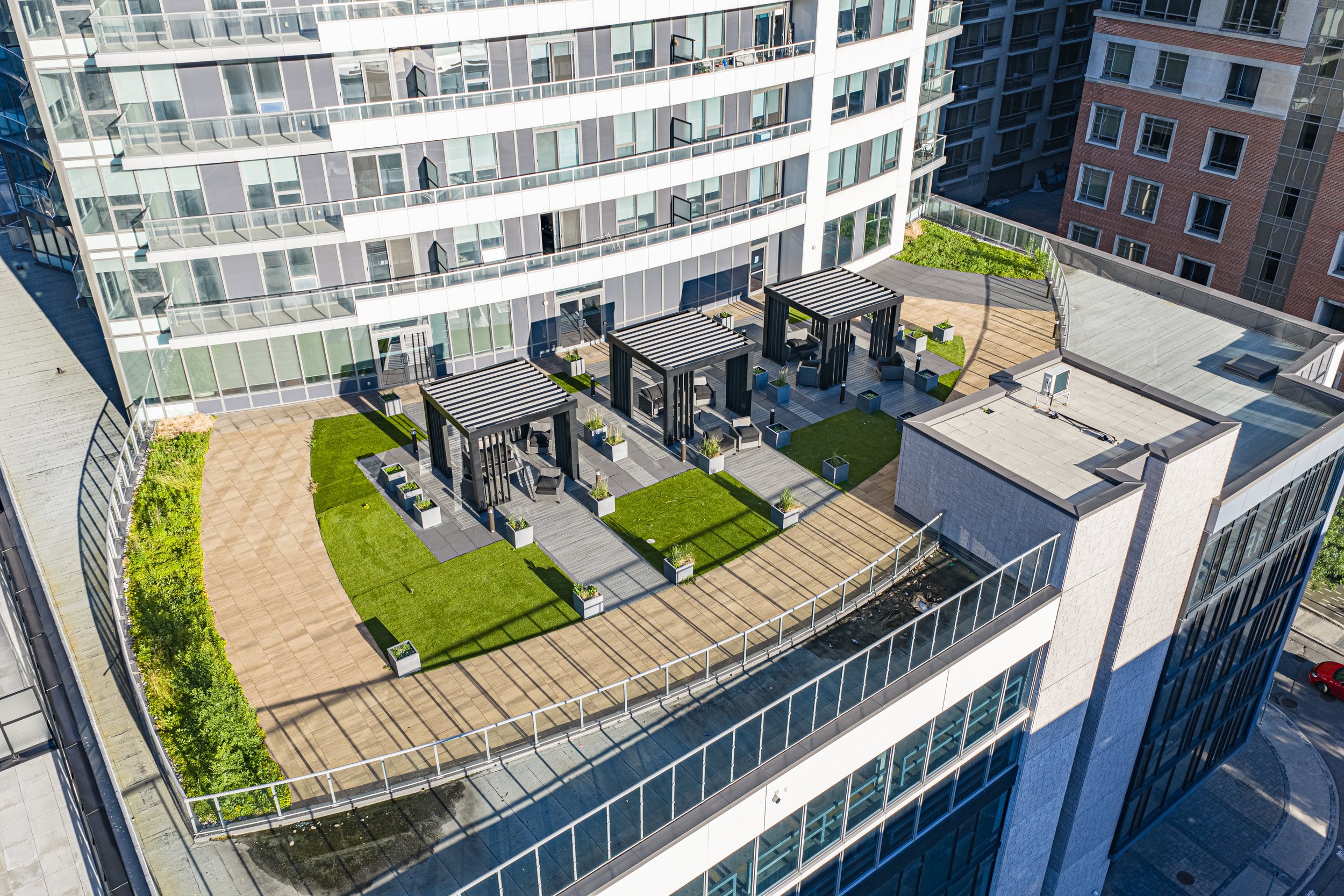 A rooftop terrace with outdoor seating, pergolas, potted plants, and a glass railing, surrounded by tall modern buildings.