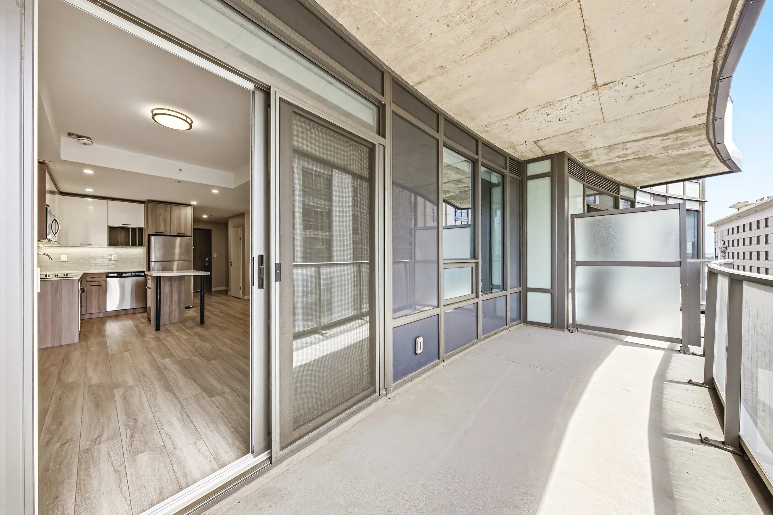 The image shows a balcony with frosted and clear glass panels, overlooking city buildings. The balcony has a concrete ceiling and a concrete floor. The interior view shows a modern kitchen with wooden cabinets, white upper cabinets, and stainless ste