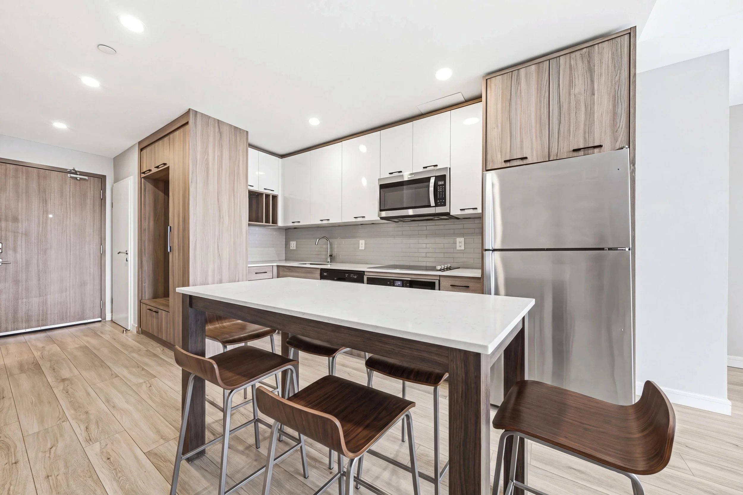 Modern kitchen with white cabinets, wood accents, stainless steel refrigerator, microwave, and seating area with wooden chairs and a white countertop.
