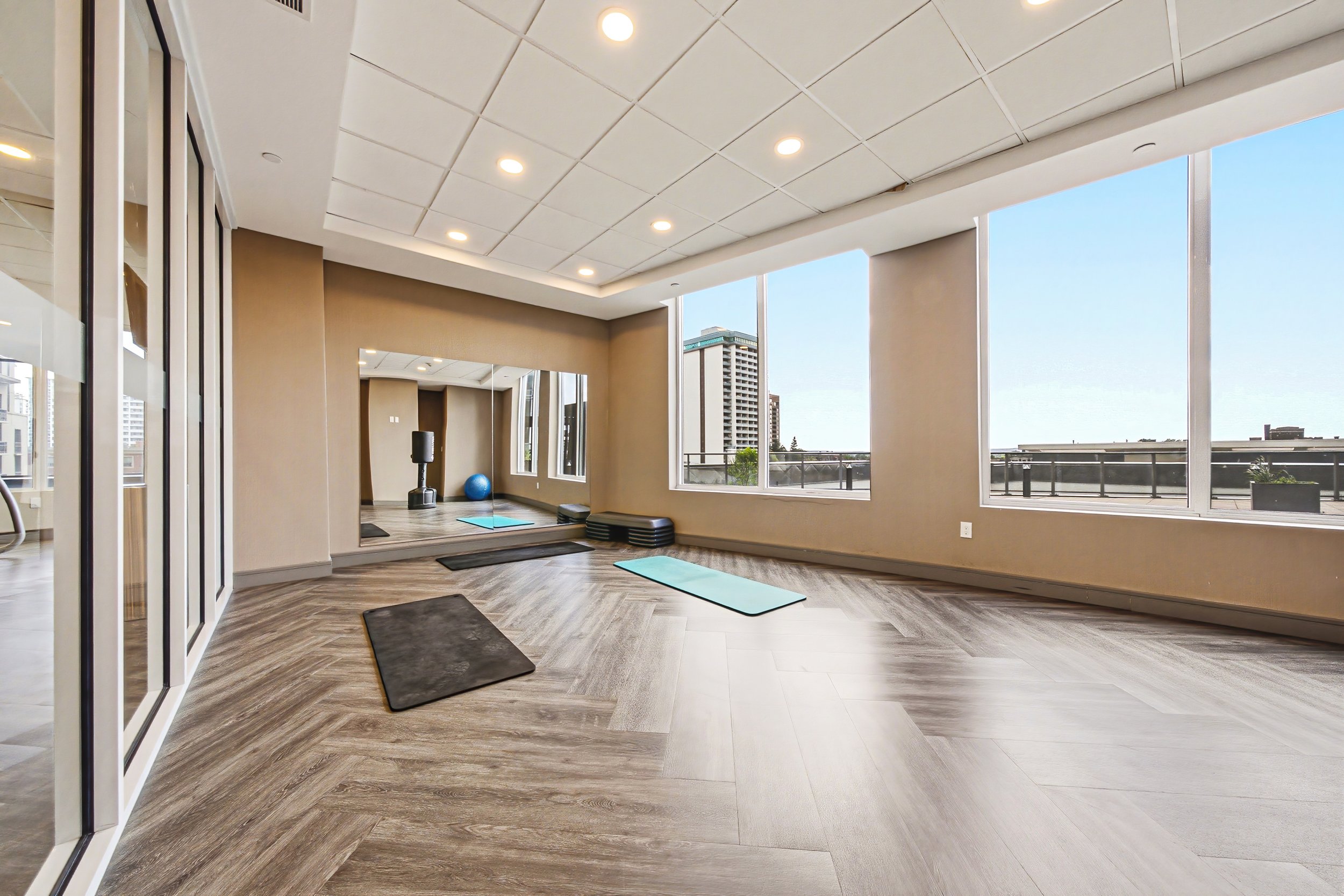 A spacious indoor exercise room with large windows showing city buildings and a blue sky. The room has wood-like flooring, recessed ceiling lights, and mirrors on one wall. There are yoga mats and workout equipment, including a punching bag and a sta