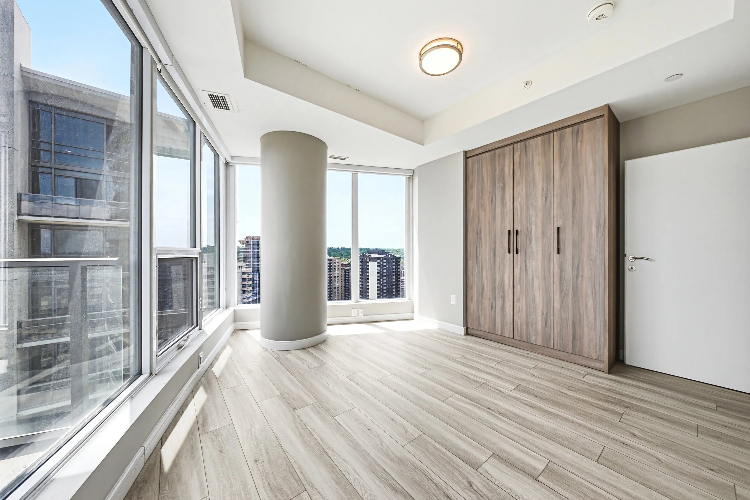 Bright, modern high-rise apartment with large windows, light wood flooring, a white ceiling with a ceiling light, a wooden closet door, and a city view outside.