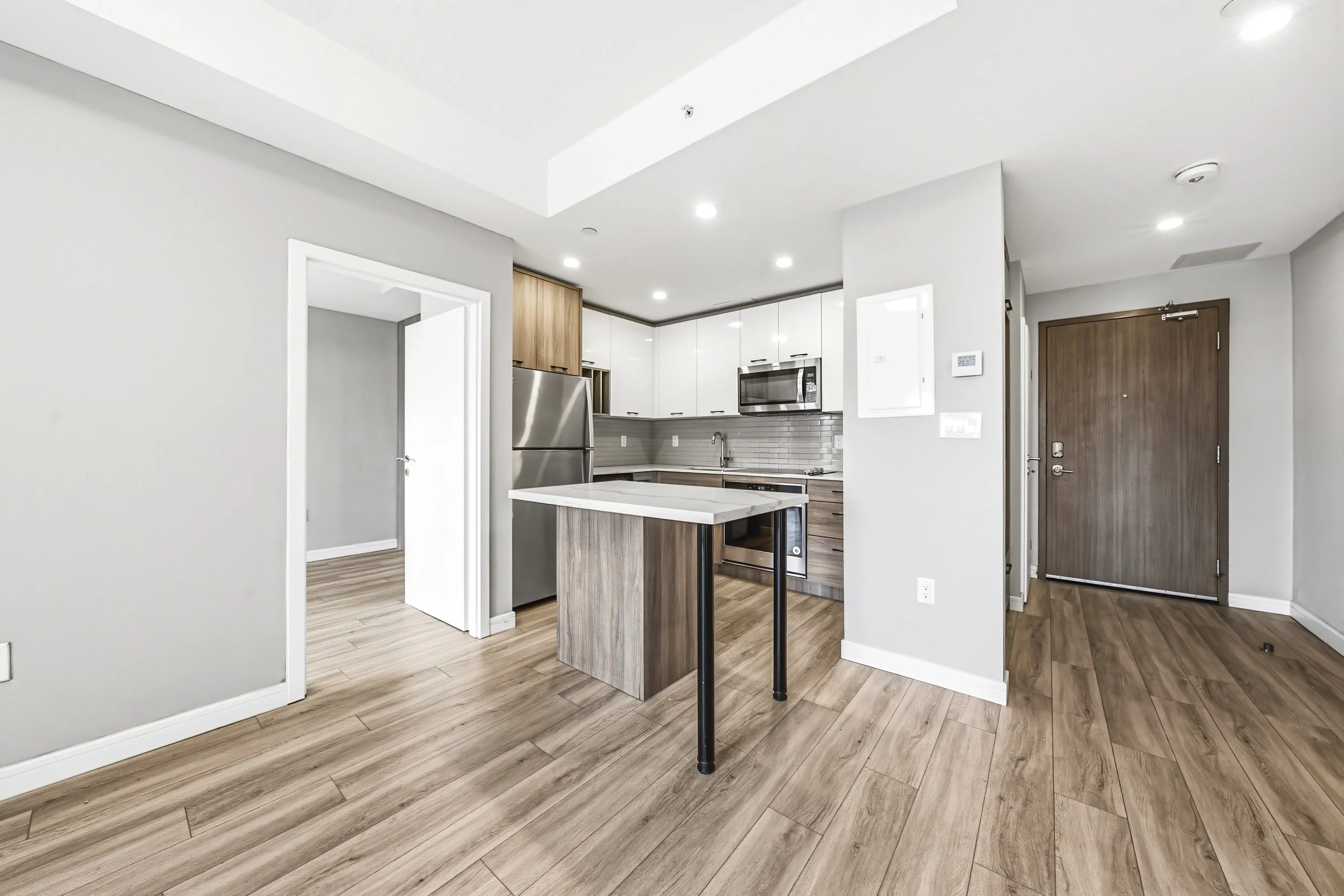 Modern kitchen with light grey walls, wooden flooring, white and wood cabinets, stainless steel appliances, and a small breakfast bar.