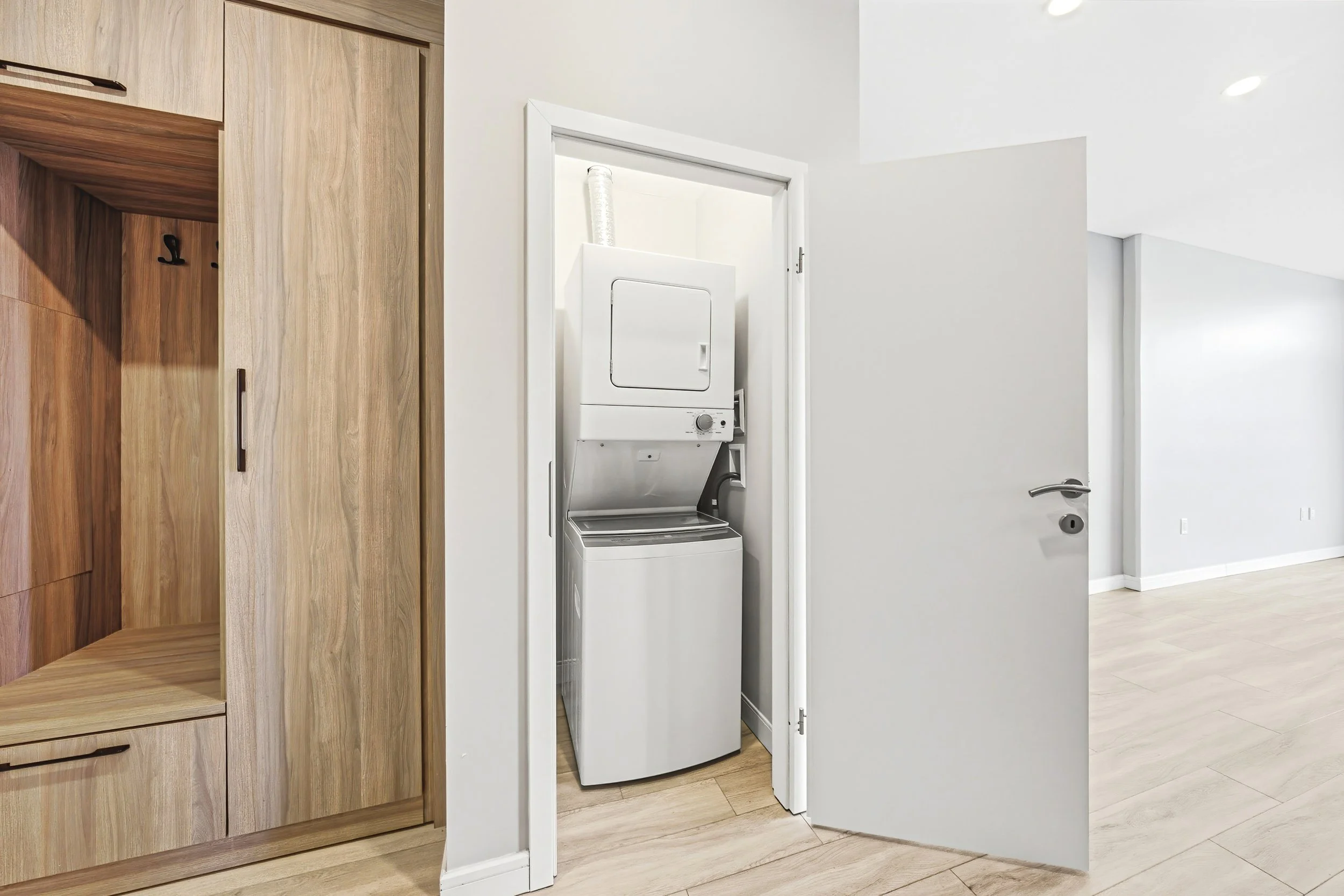 A laundry area with a white stackable washer and dryer behind a partially open white door. The room has wood cabinetry and light-colored flooring.
