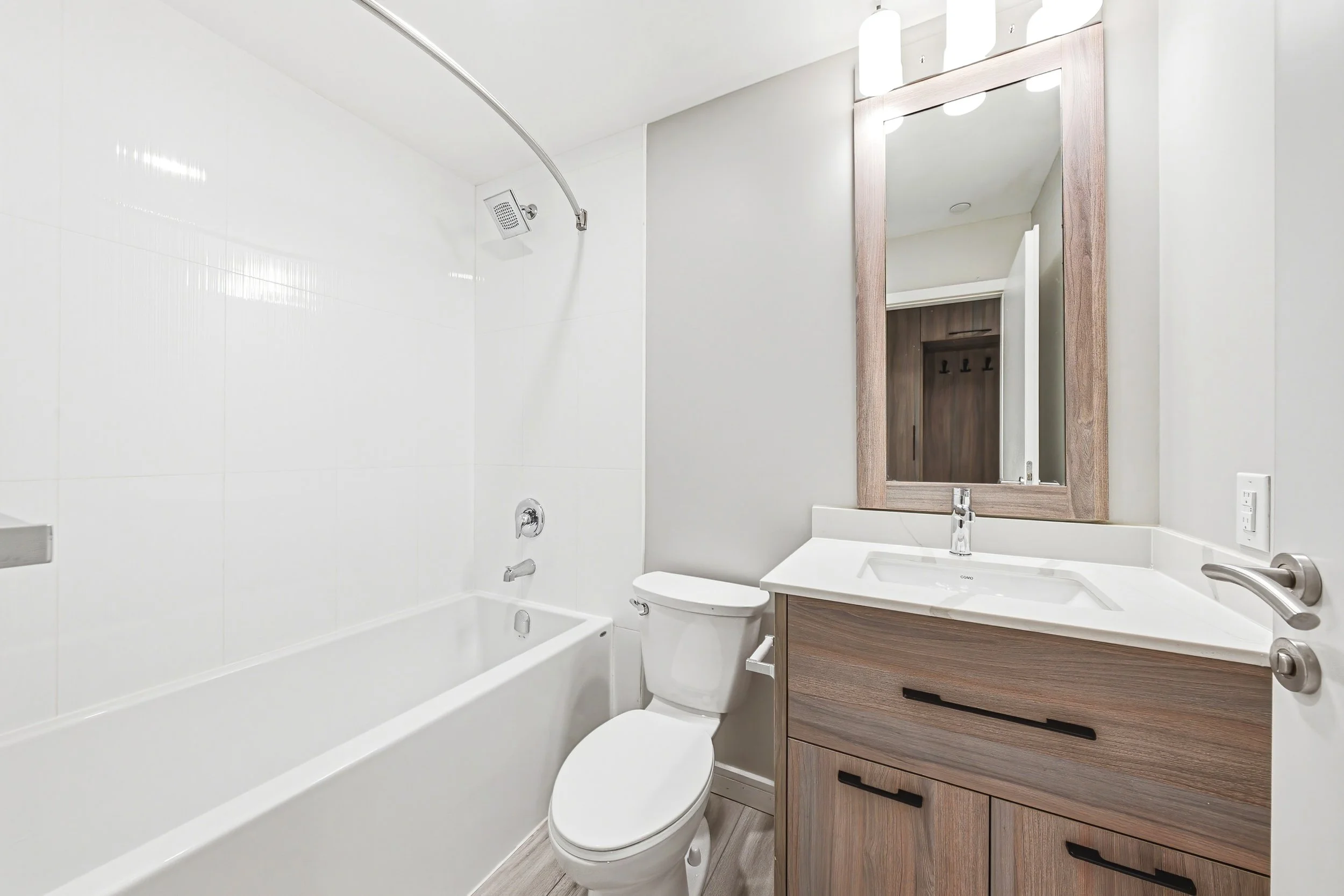 A modern bathroom featuring a white bathtub with a shower head and a white toilet. Next to the toilet is a wooden vanity with a sink, a large mirror, and overhead lighting. The walls are plain and light-colored, creating a clean and minimalist look.