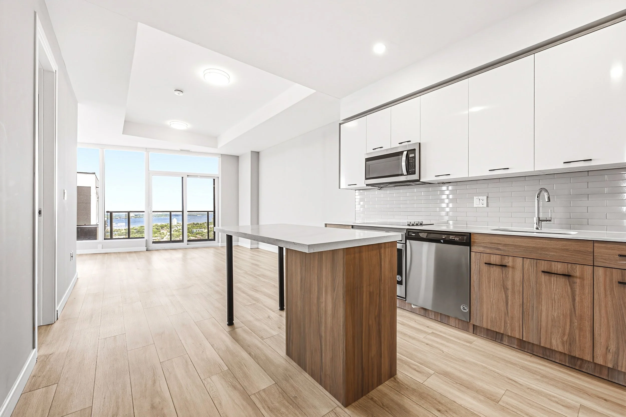 Modern kitchen with white cabinetry, wooden lower cabinets, a white countertop island, stainless steel appliances, and a view of a balcony and water outside.