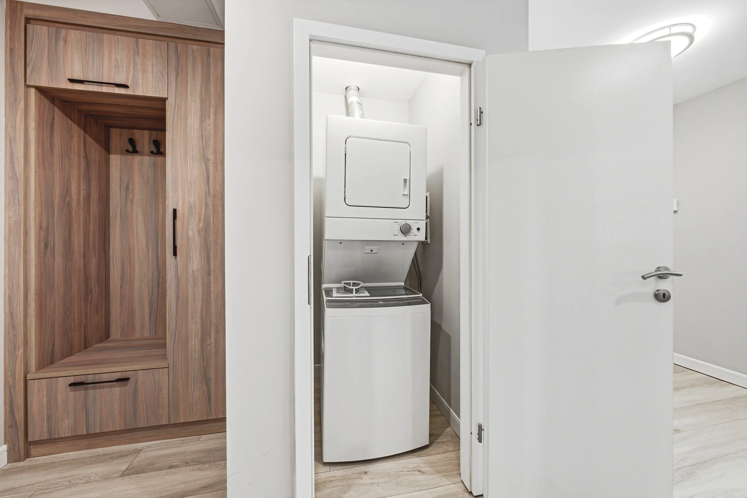 A laundry area with a stacked washer and dryer behind a door, a wooden closet on the left, white walls, and light wood flooring.
