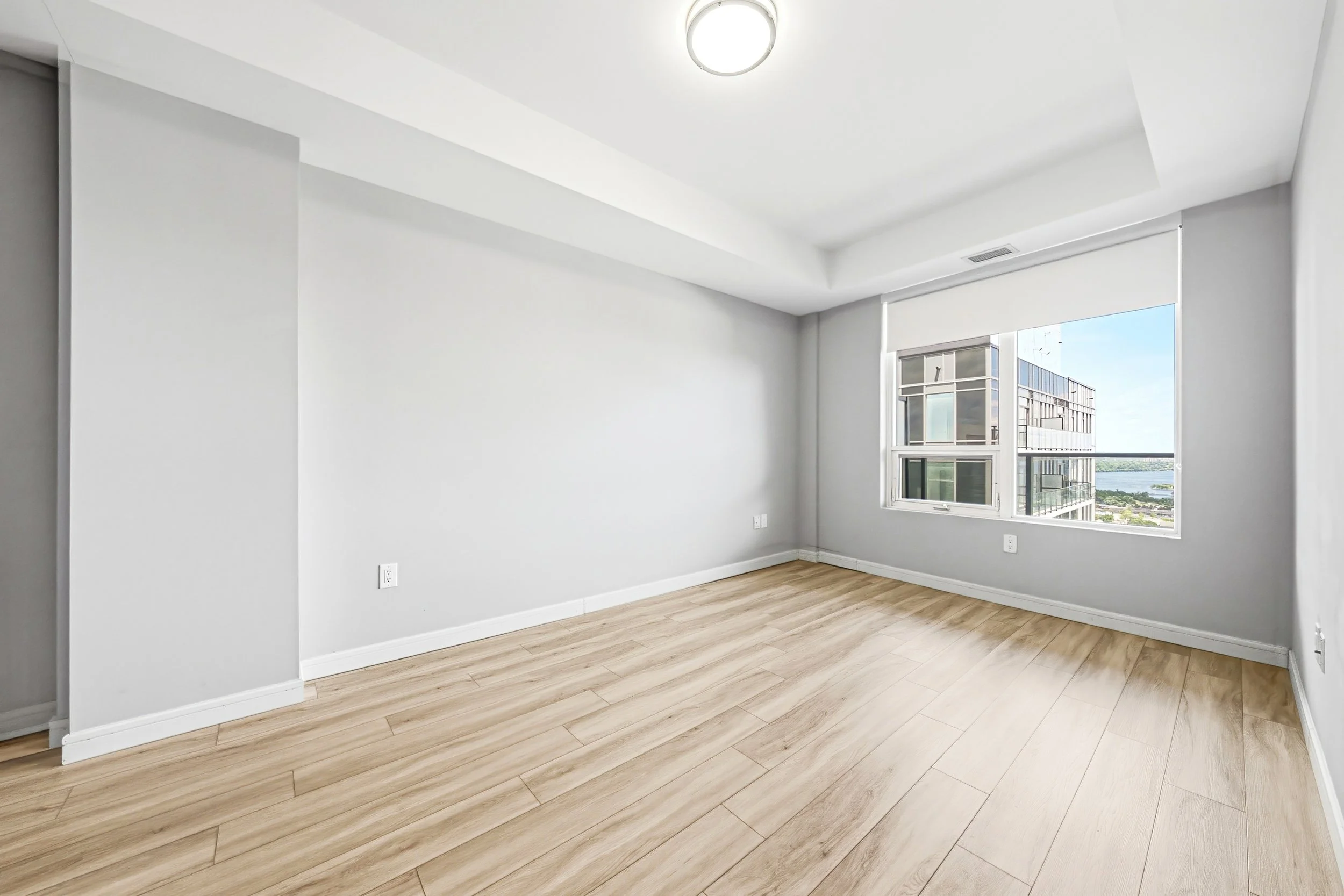 Empty modern room with white walls, large window with a view of a cityscape, and light wood flooring.