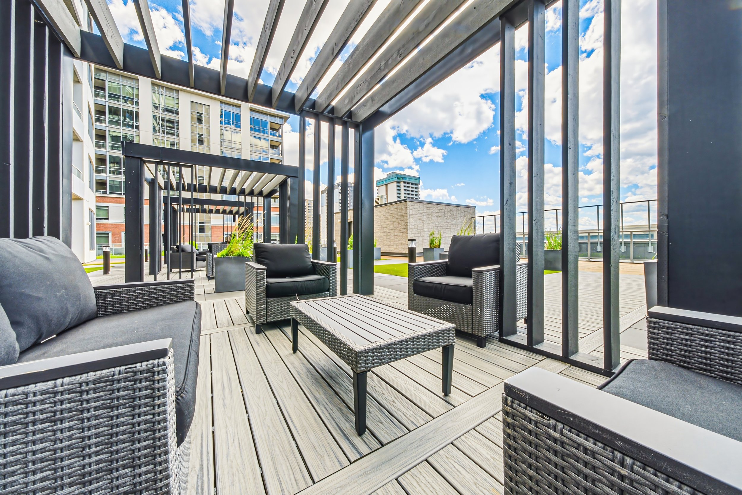 Rooftop terrace with outdoor seating, including wicker sofas and chairs, surrounded by metal slats and planters, overlooking city buildings under a partly cloudy sky.