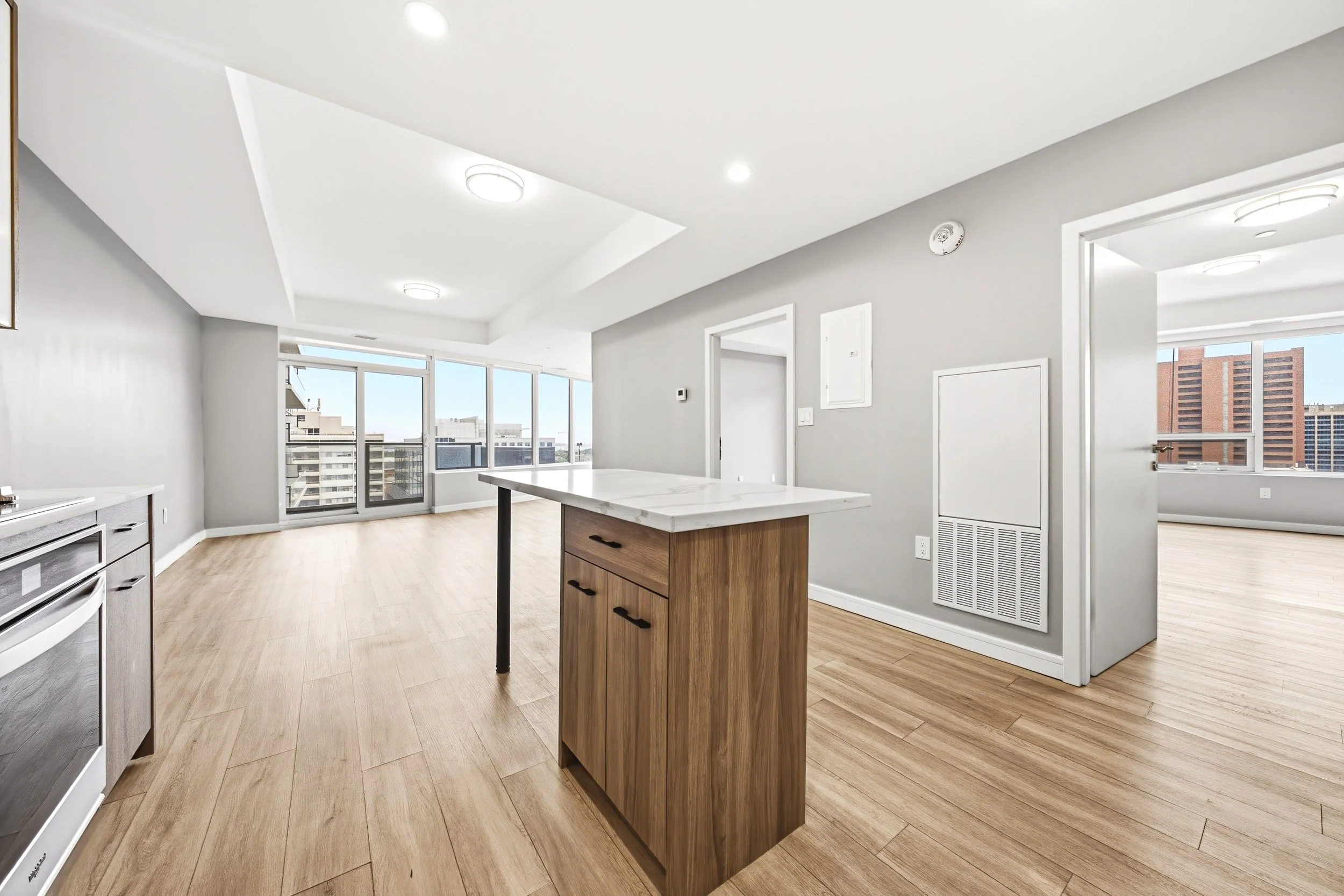Modern, empty kitchen and living area with large windows, light wood flooring, white and gray walls, central wooden kitchen island, and cityscape view.