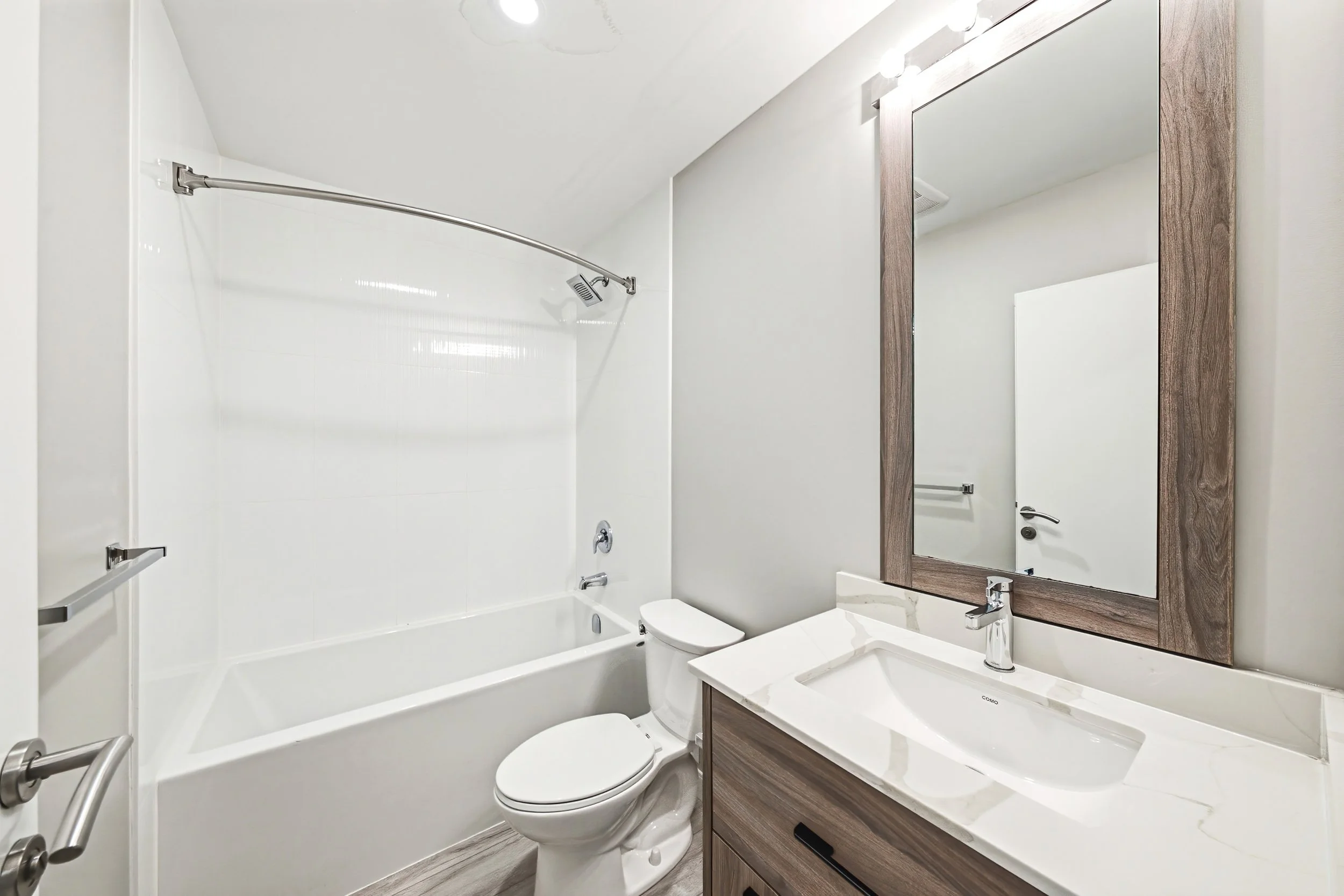 Modern bathroom with white bathtub, showerhead, wooden framed mirror, white toilet, and white marble countertop.