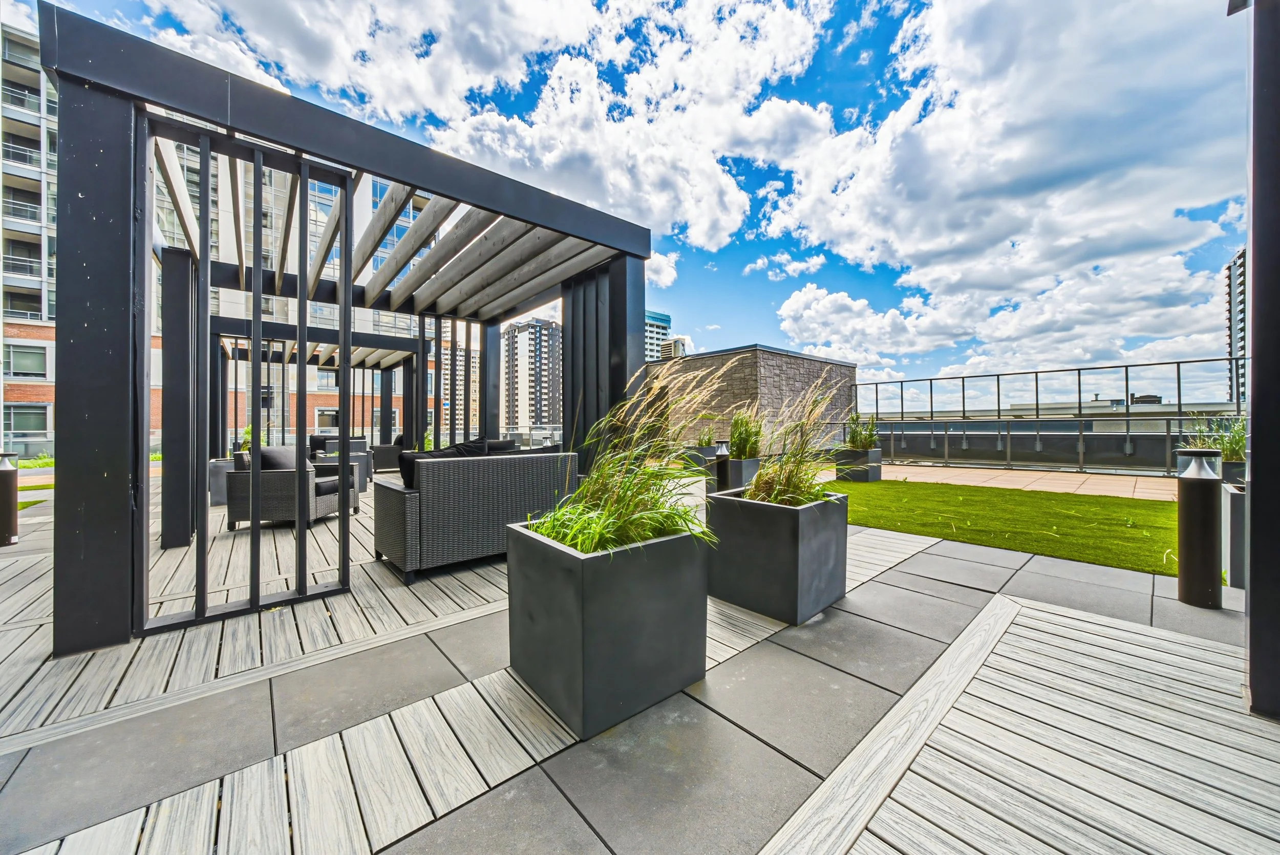Rooftop terrace with outdoor furniture, planters, and a partially covered seating area, with city buildings and a partly cloudy sky in the background.
