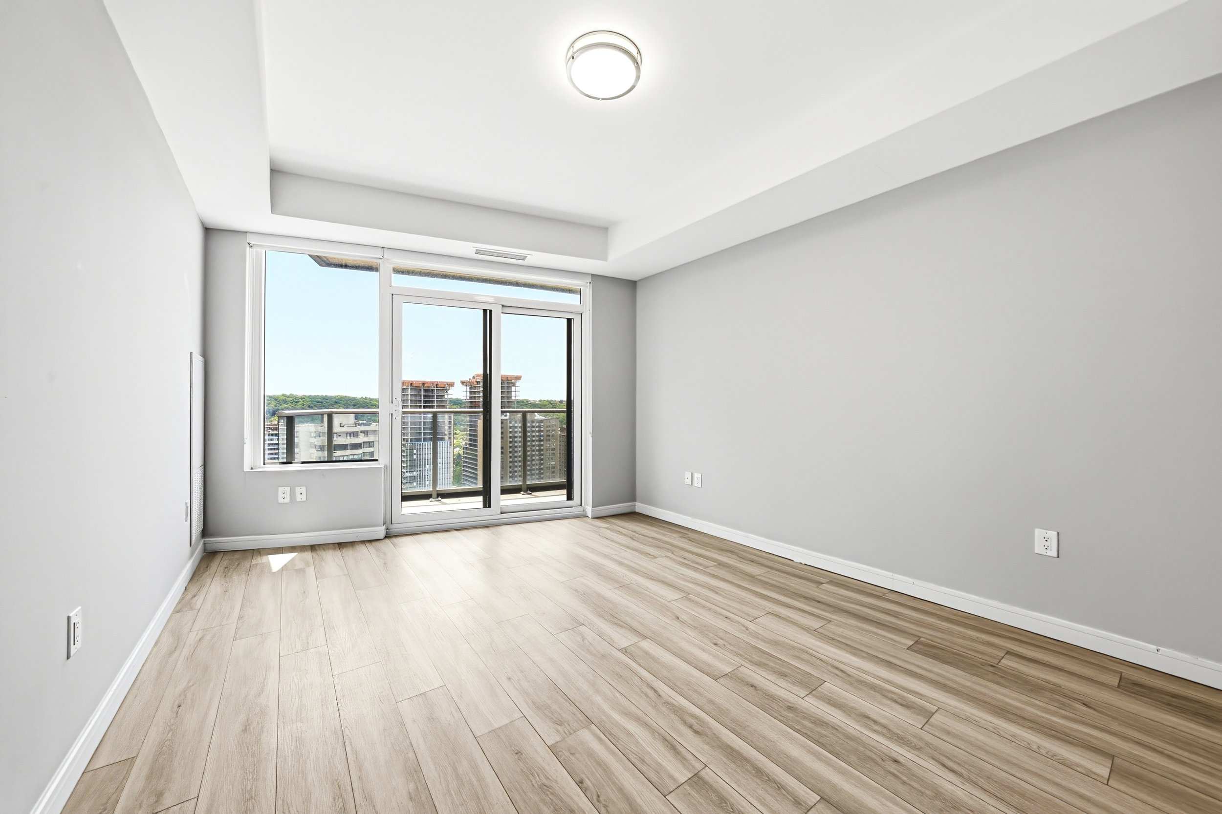 Empty room with light-colored wooden floors, white walls, a large window with a balcony, and a ceiling light fixture.