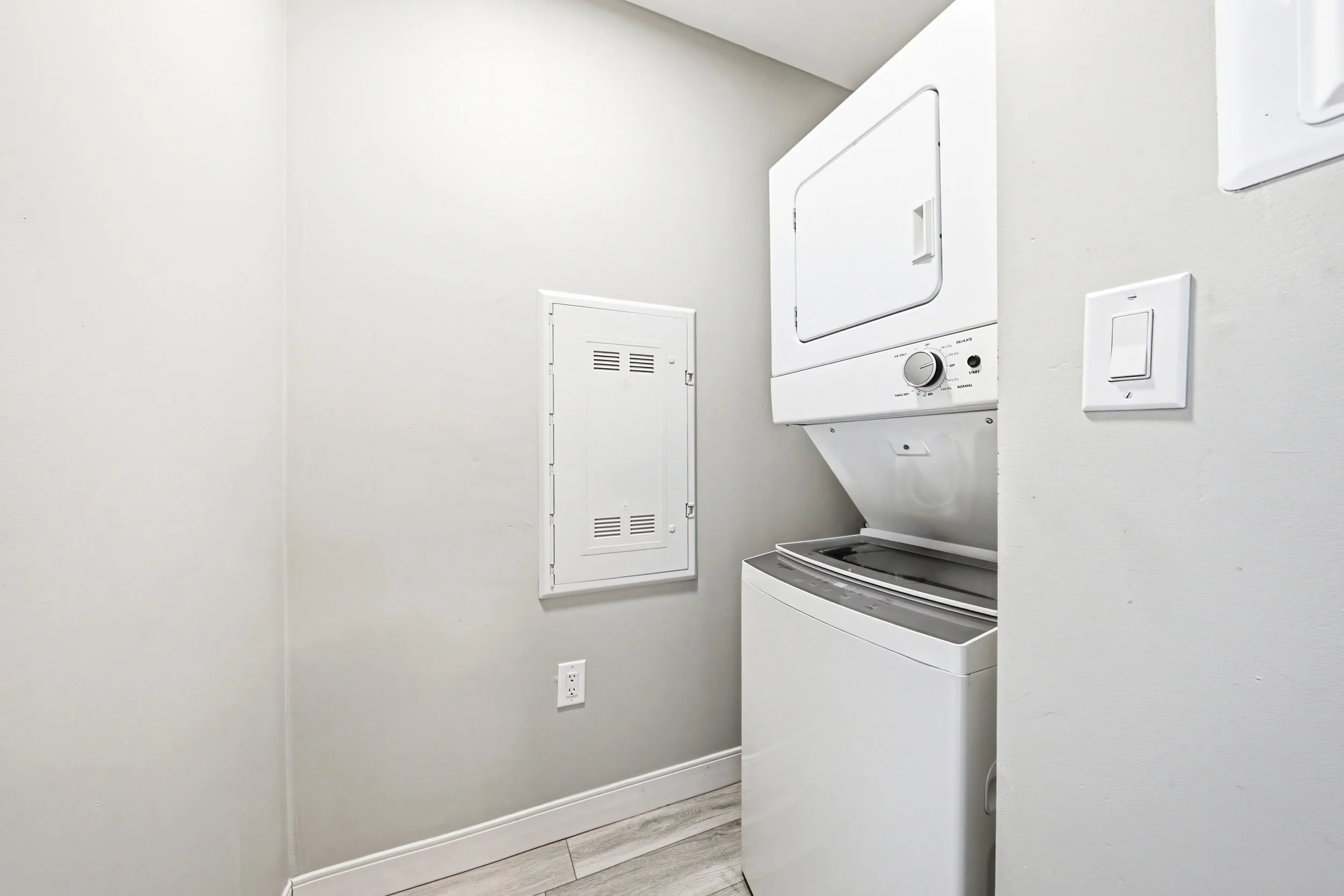 A laundry area with a stacked washer and dryer, white walls, a small white door, a light switch, power outlet, and light wood flooring.