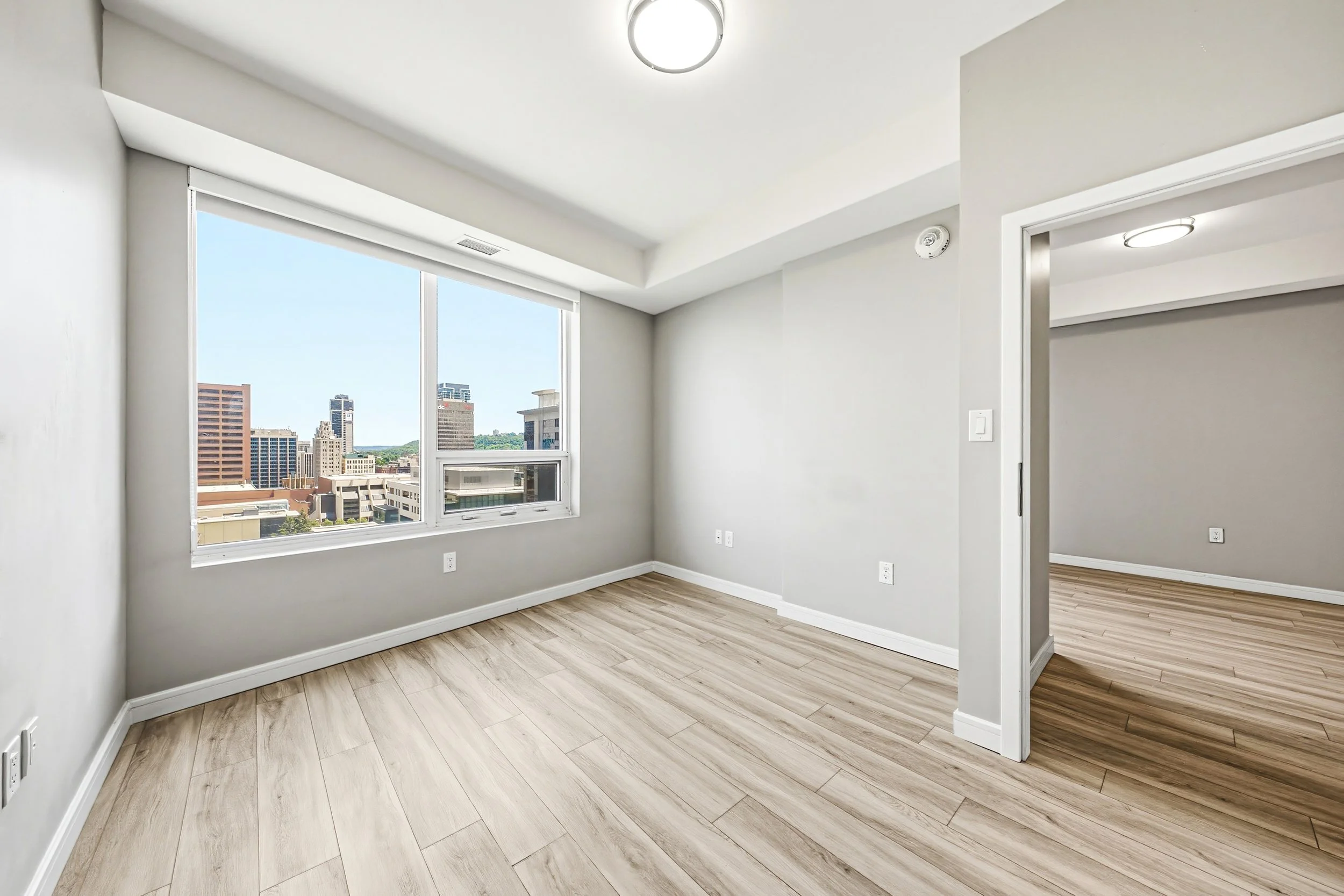 Empty modern apartment room with large window showing city skyline, light wood floors, white walls, and ceiling lighting.