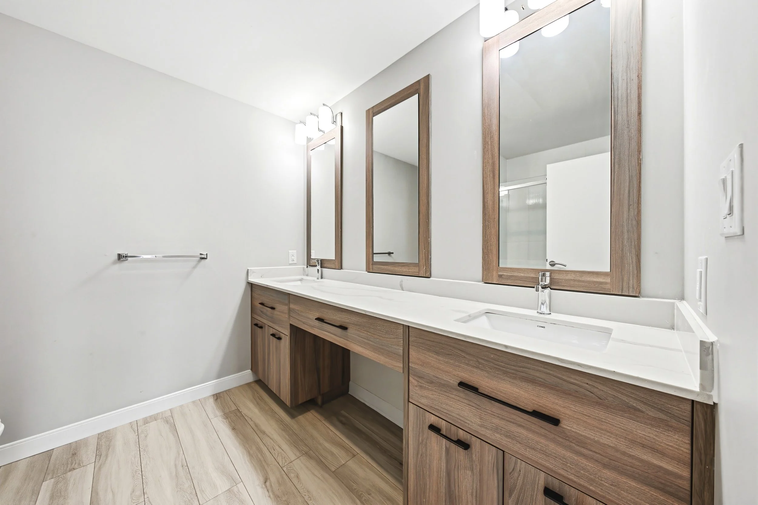 Modern bathroom vanity with two mirrors, wooden cabinets, a white countertop, and a single sink. Light fixtures are mounted above the mirrors, and there is a towel rack on the wall.
