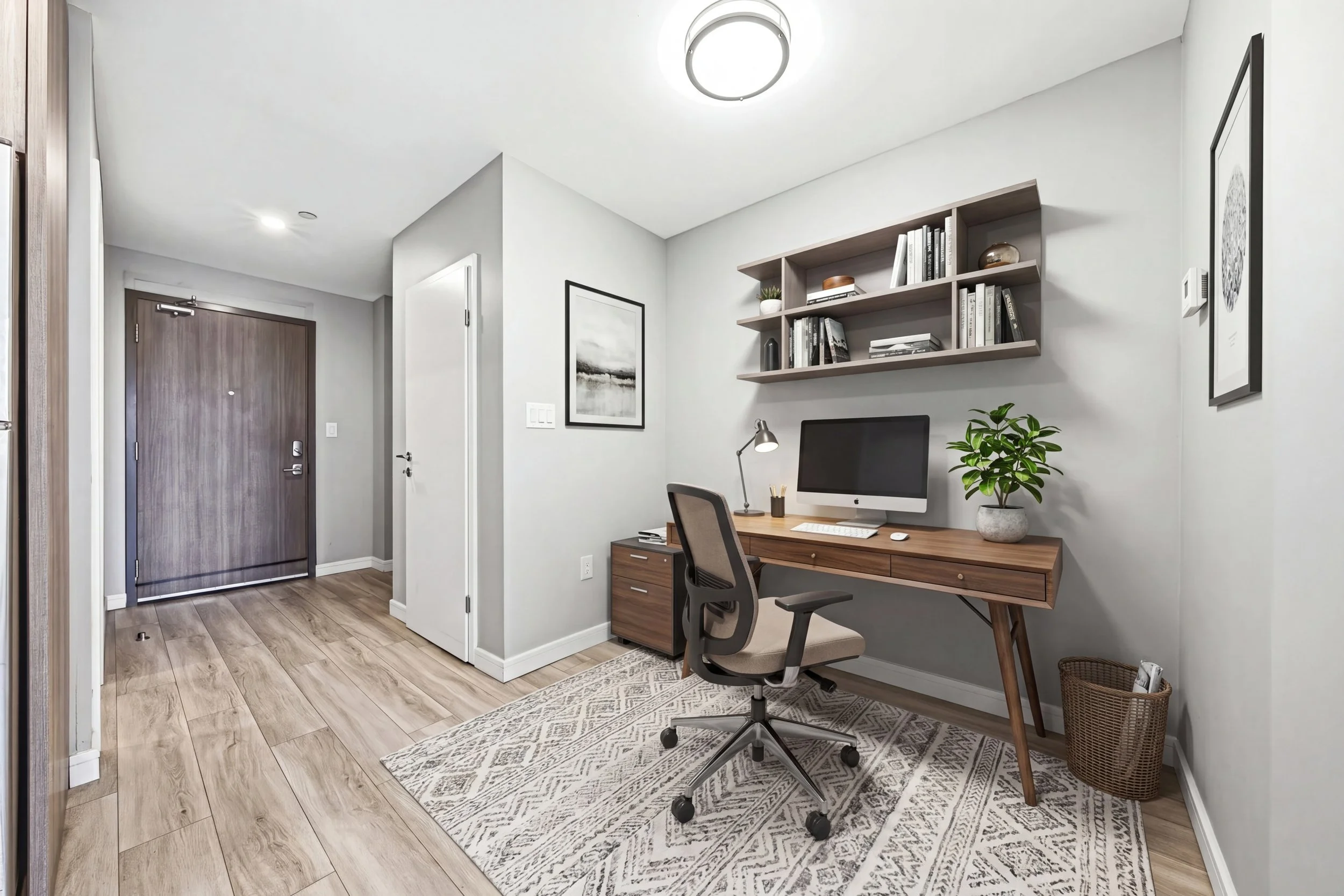 A modern home office with a wooden desk holding a computer monitor, desk lamp, and a potted plant. A black office chair is in front of the desk. A bookshelf with books and decor is mounted on the white wall. There is a patterned rug on the light wood