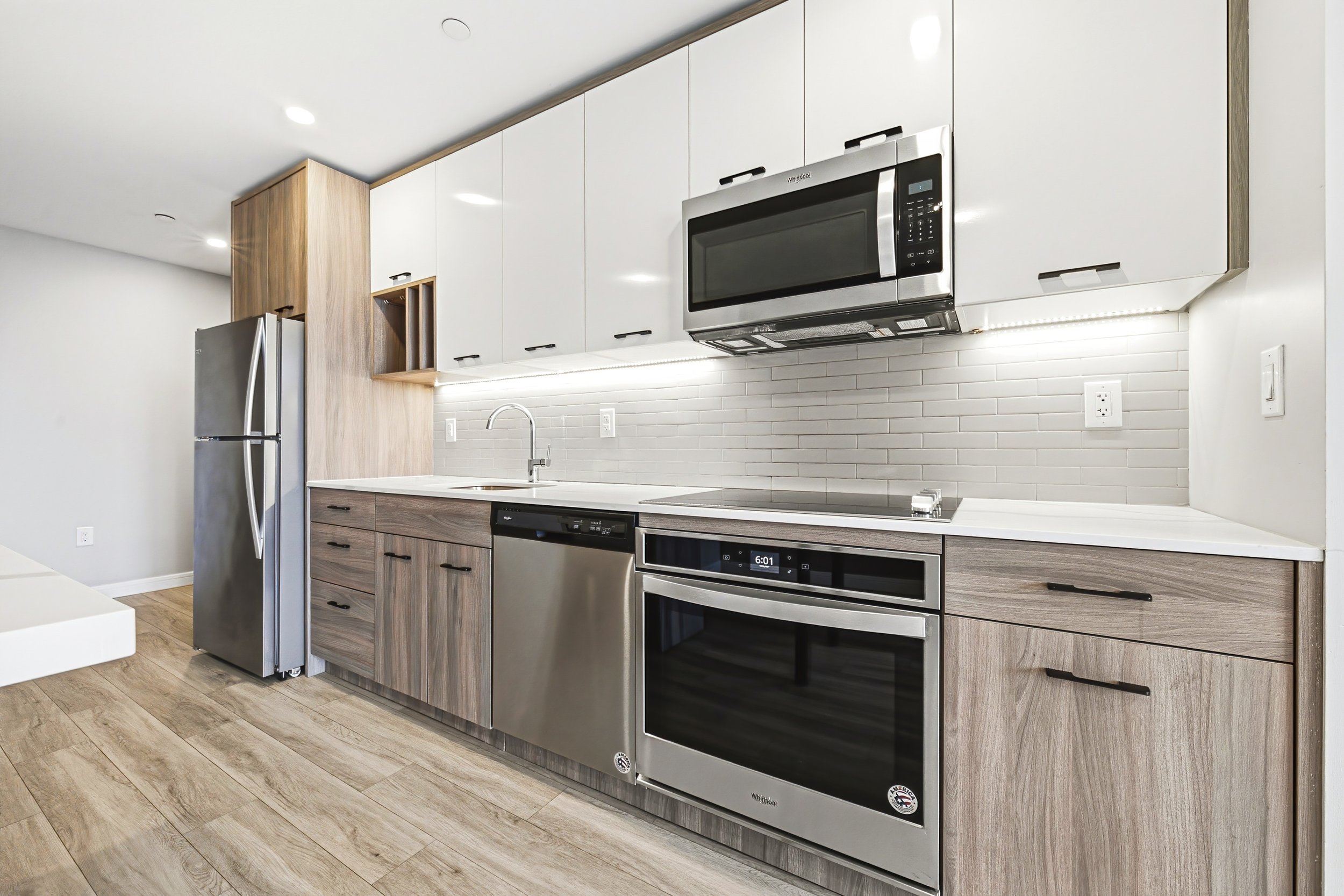 Modern kitchen with white upper cabinets, wood lower cabinets, stainless steel appliances including a microwave and refrigerator, white tile backsplash, and wood-look flooring.