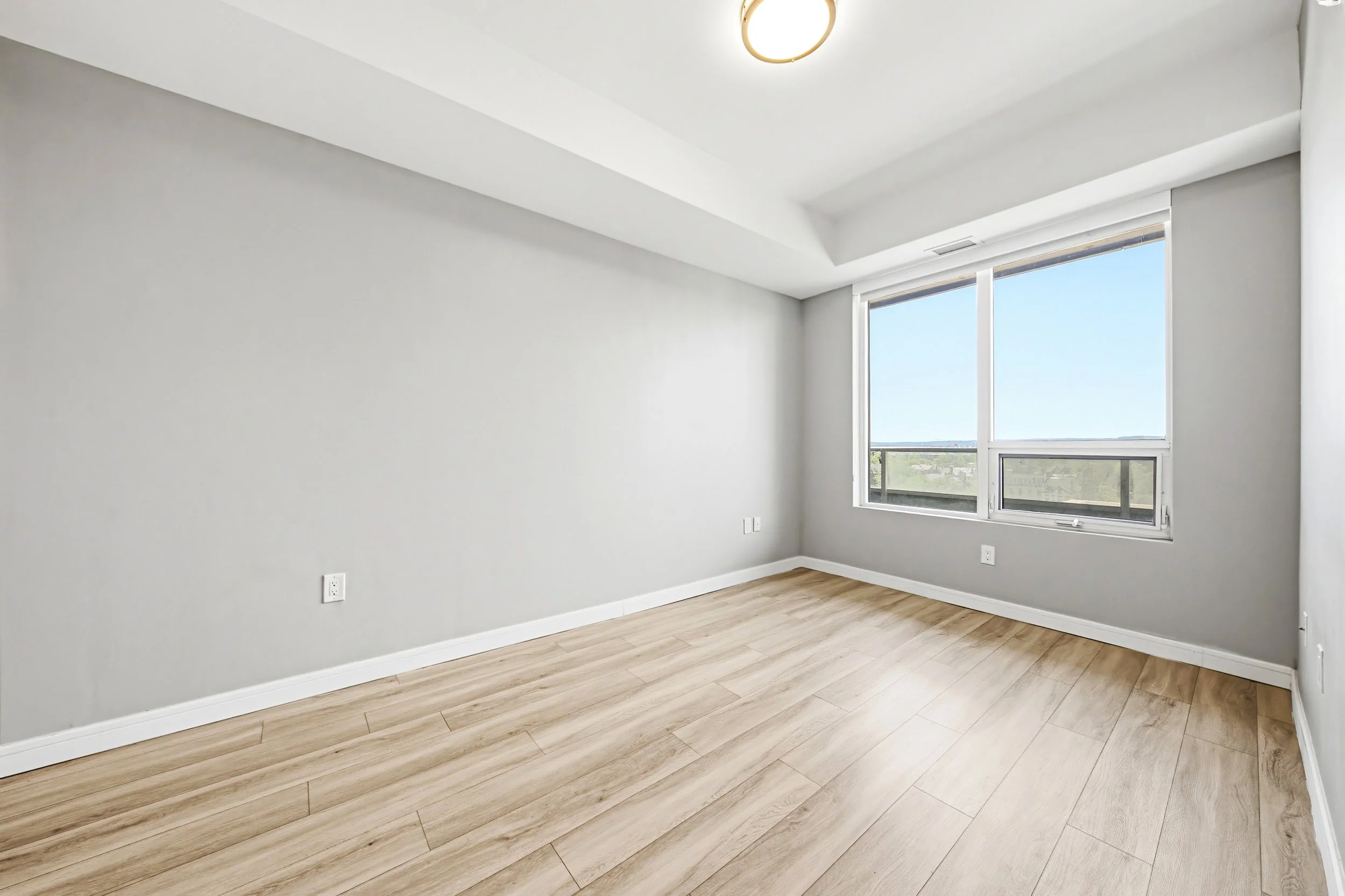 Empty room with light wooden flooring, white walls, and large windows with a view of a clear sky.
