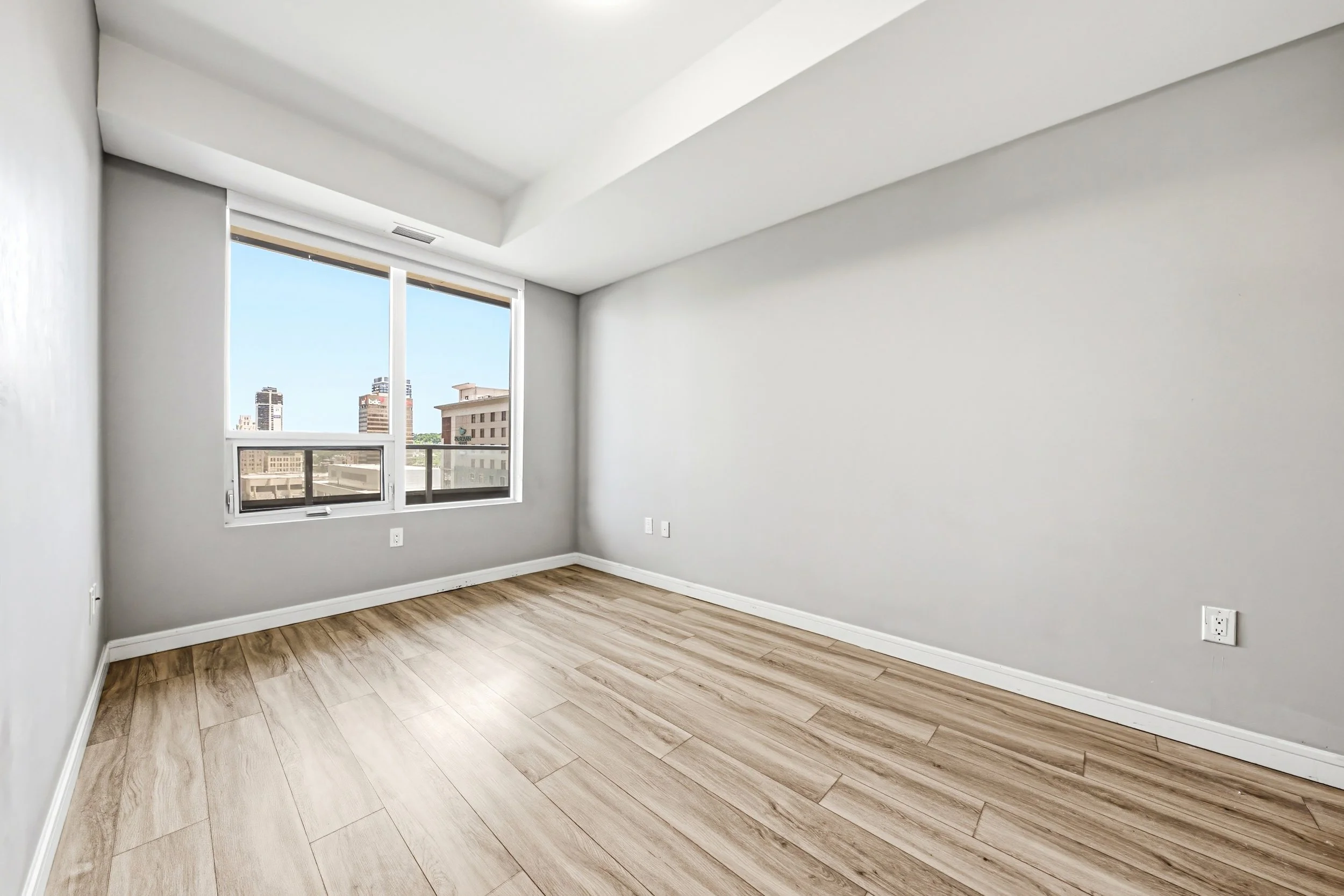 An empty room with white walls, large window showing city buildings, and light wood flooring.
