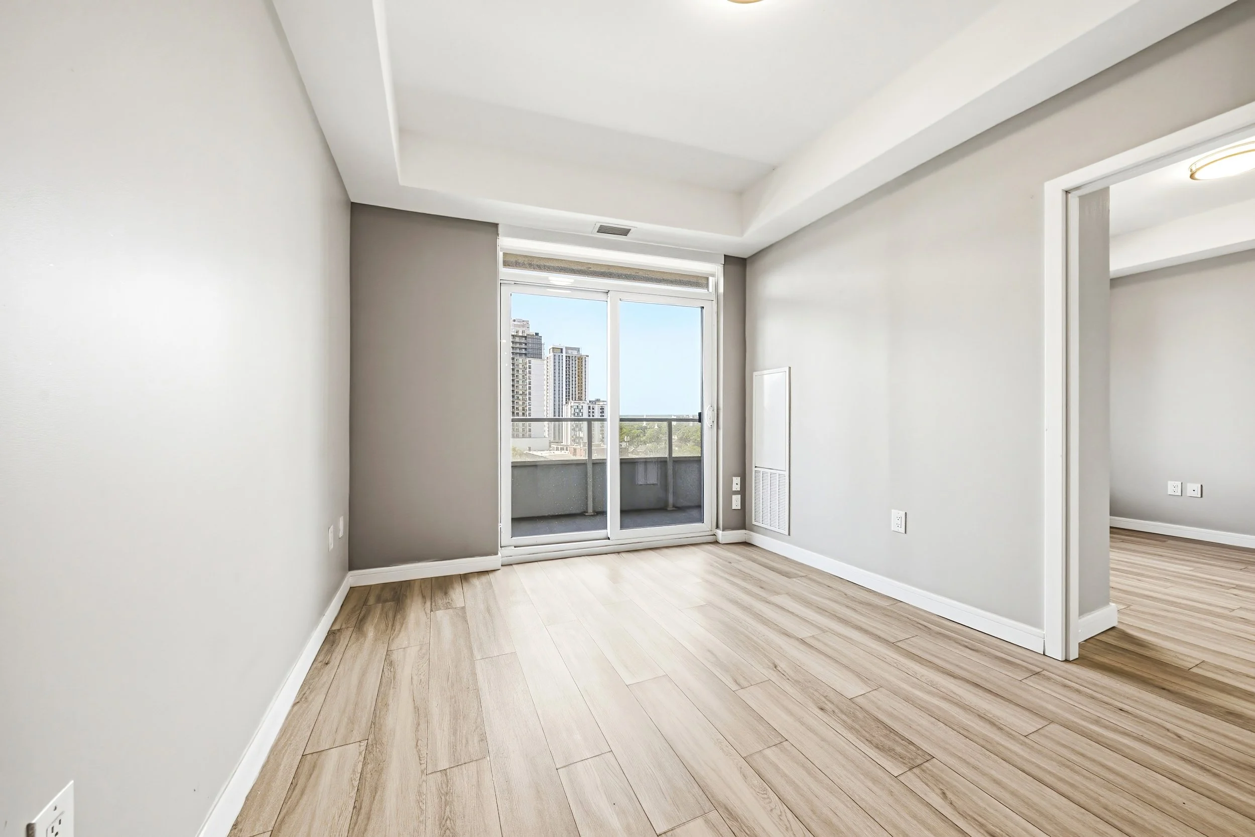 Empty room with light wood flooring, white walls, large window with city view, and a balcony door.
