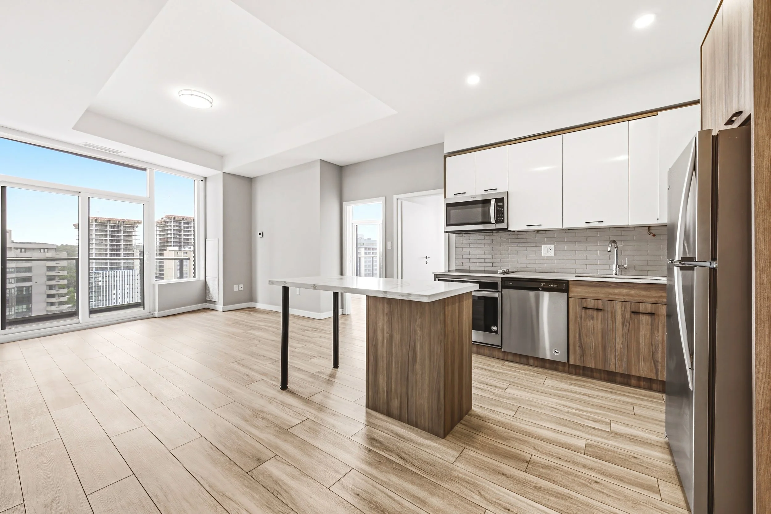Modern kitchen with white cabinets, stainless steel appliances, a marble kitchen island, light wood floors, and large windows with city views.