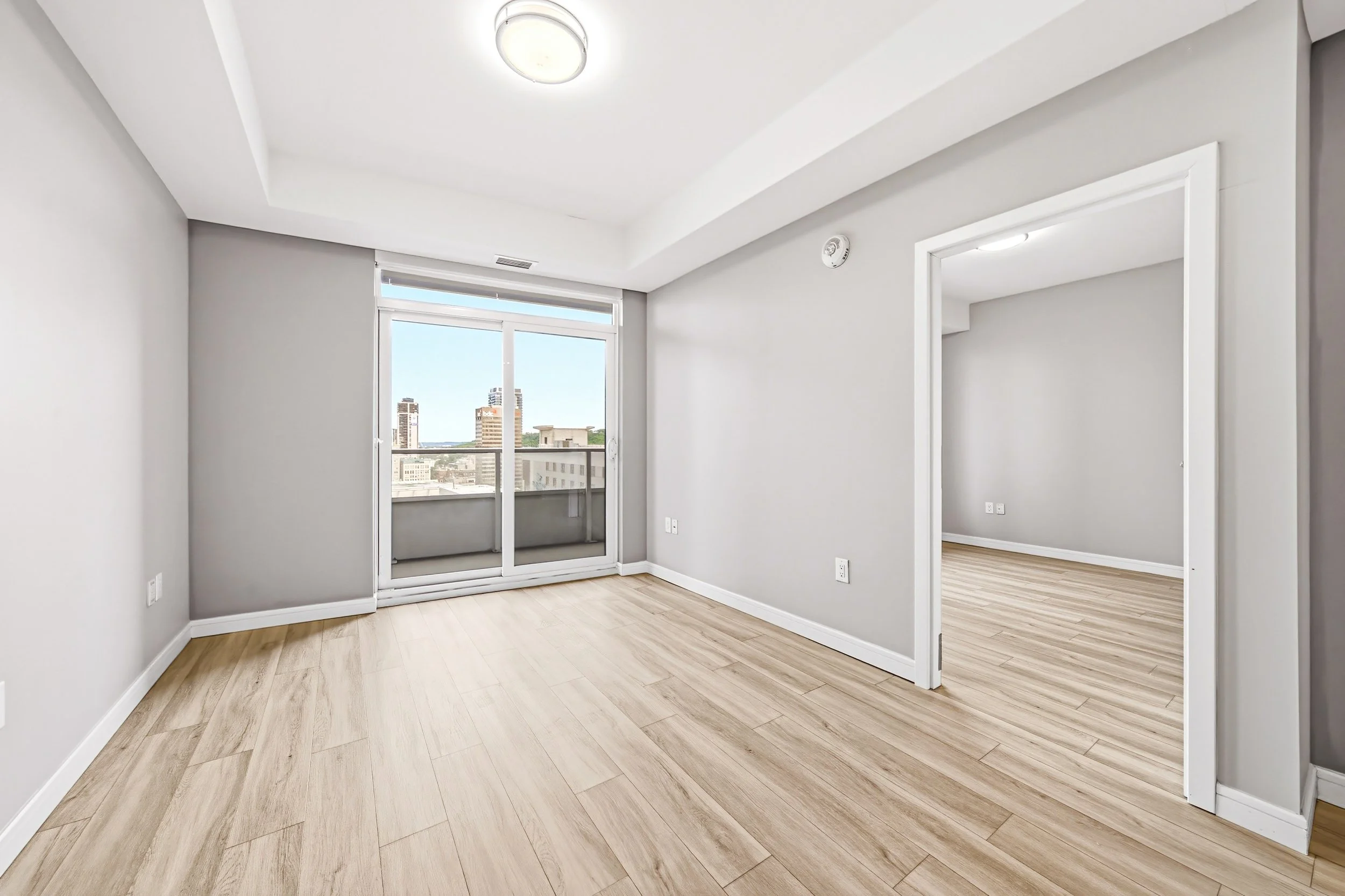 Empty living room with large windows, view of city buildings, light wood flooring, gray walls, and white trim.