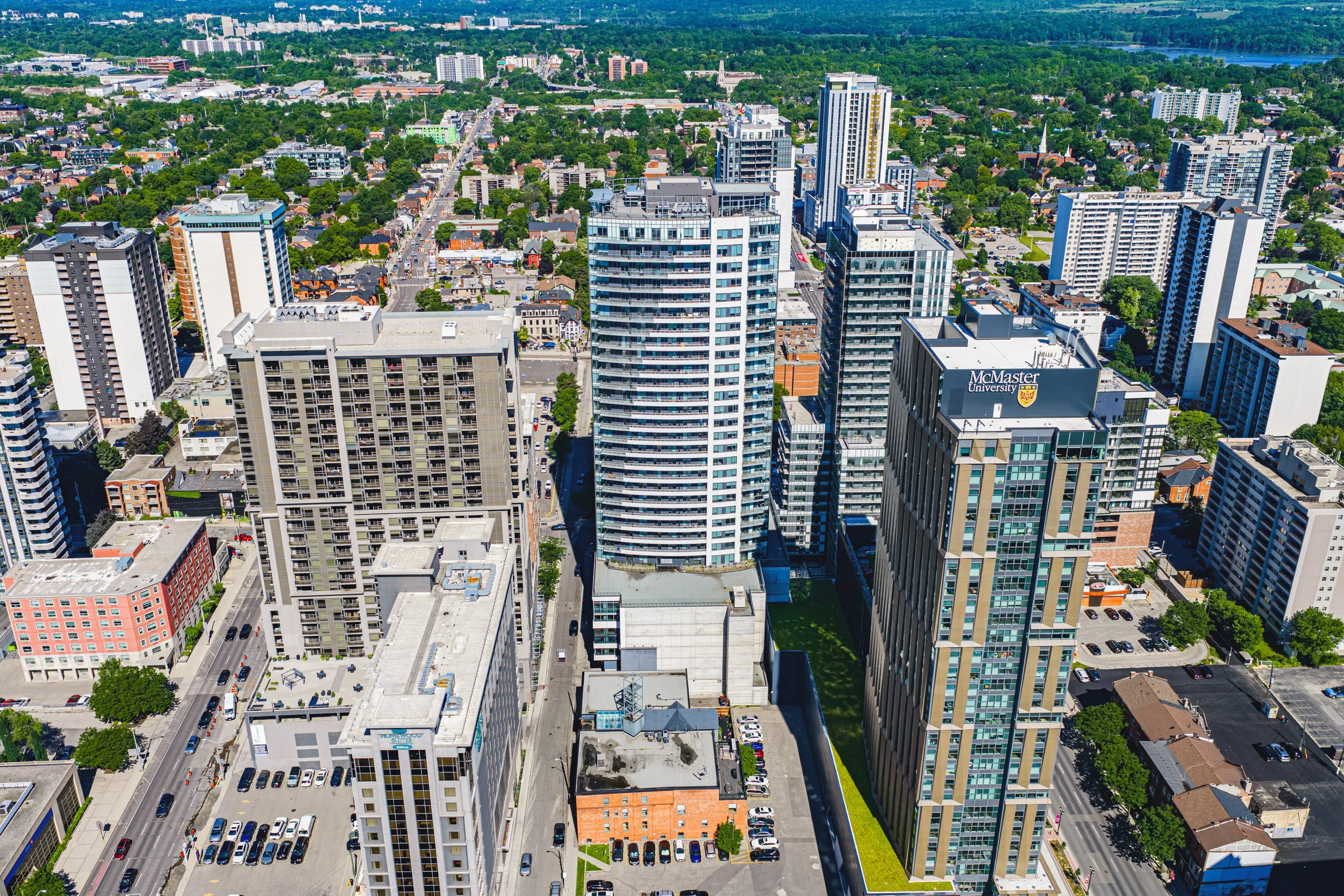 Aerial view of a city skyline with tall modern buildings, streets, parking lots, and green trees, with a distant river in the background.