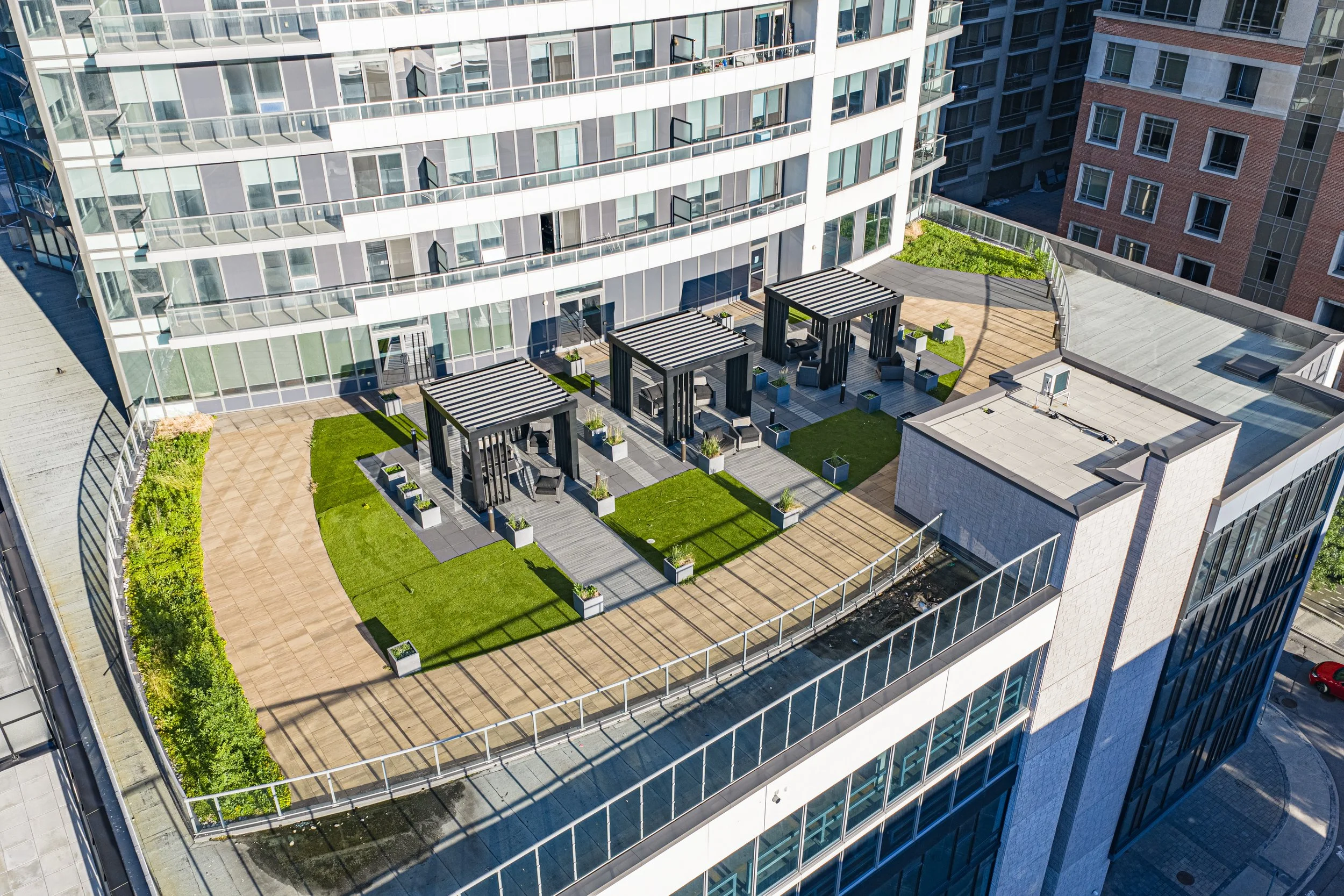 Aerial view of a modern high-rise apartment building with a rooftop terrace featuring seating areas, pergolas, potted plants, and green spaces, surrounded by other tall buildings.