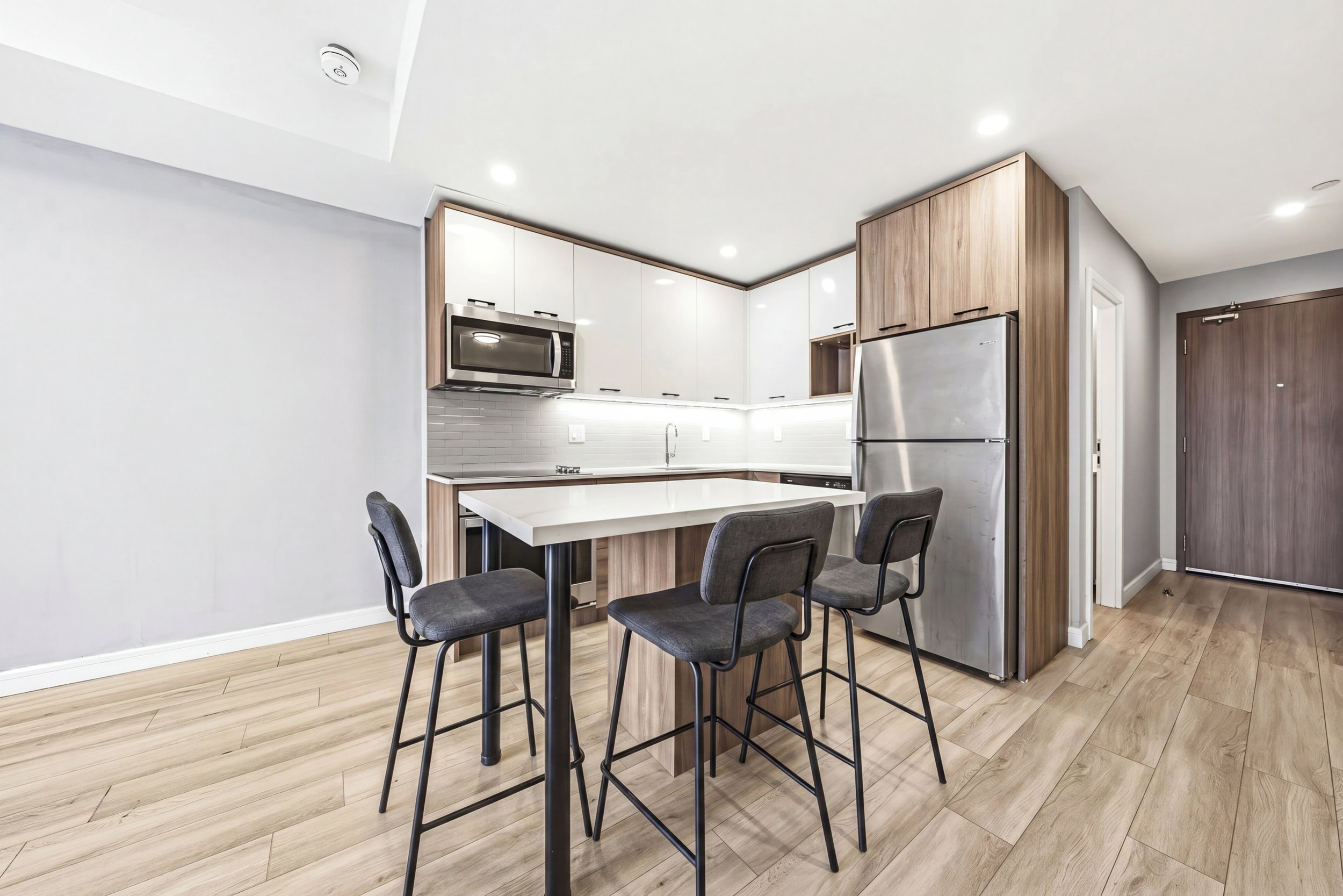 Modern kitchen with white and wooden cabinets, stainless steel microwave and refrigerator, gray chairs around a white kitchen island, and light wood flooring.