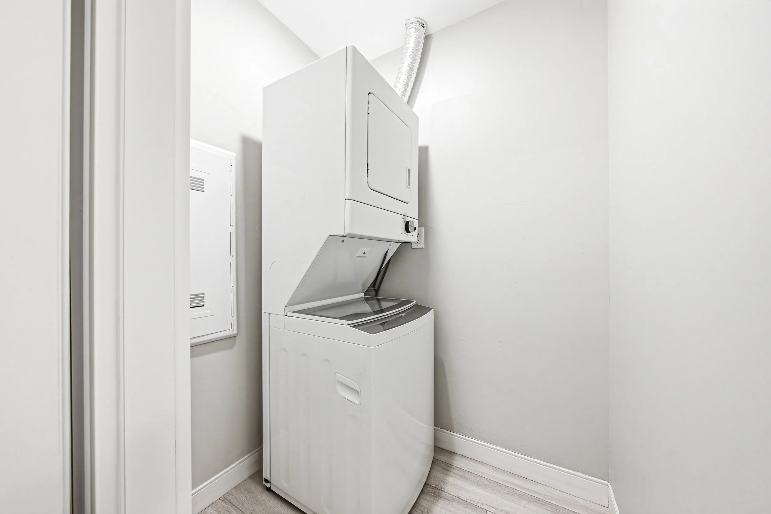 A white stacked washer and dryer unit in a small laundry room with white walls and light-colored wood flooring.